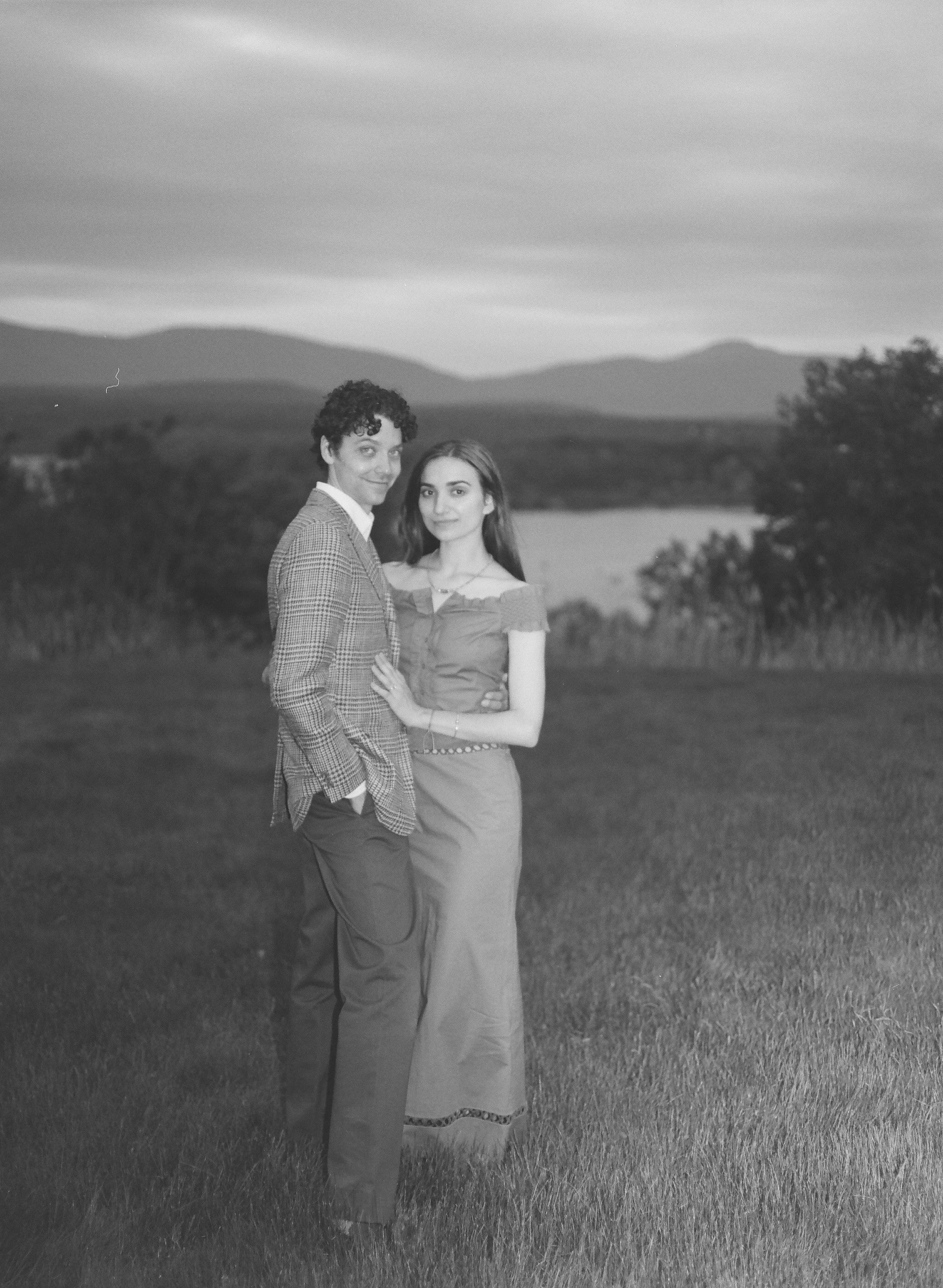 engaged couple stand in the grass looking at camera with the hudson river and catskill mountains behind them during their engagement photo shoot at blithewood gardens