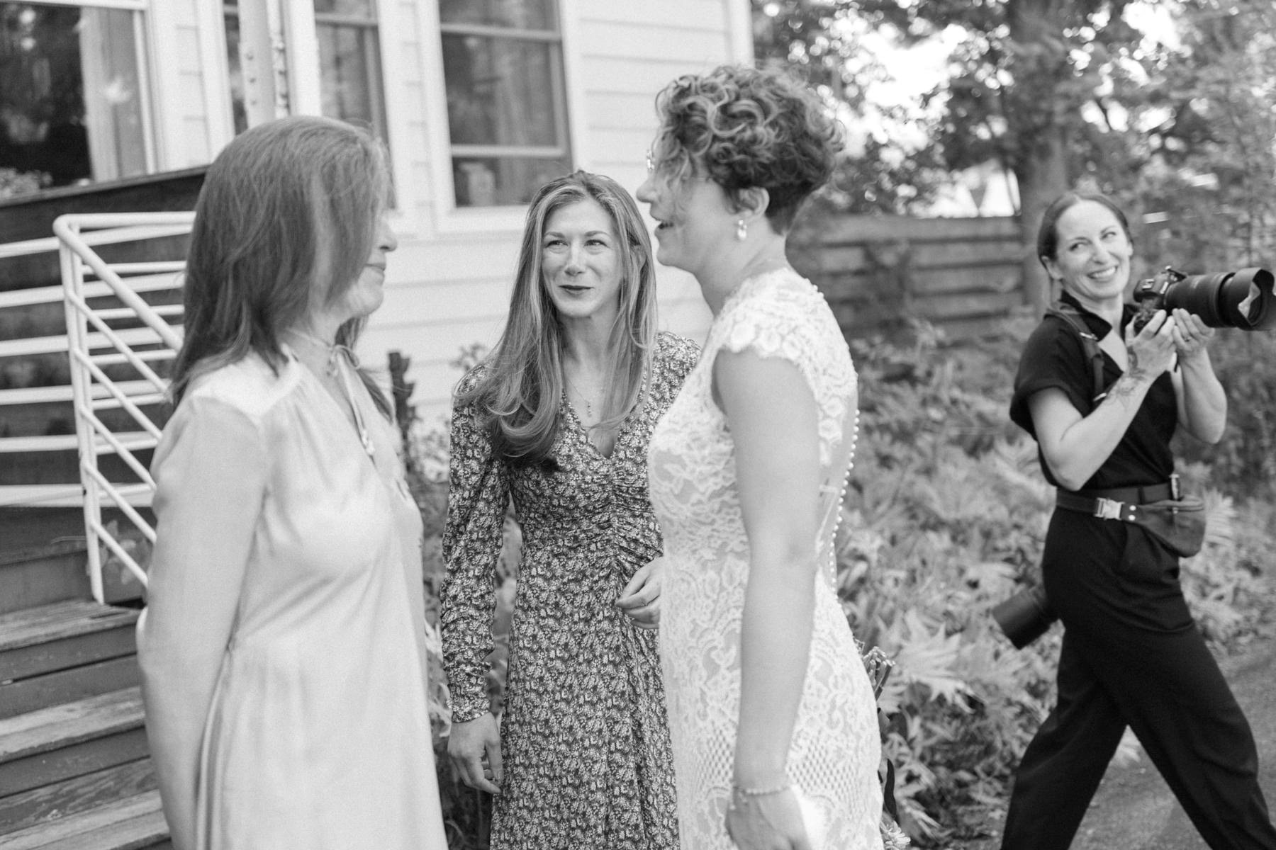 Four women, including one bride in a wedding dress, interacting outdoors with Caitlinn Mahar-Daniels, hudson valley wedding photographer, taking pictures in the background.