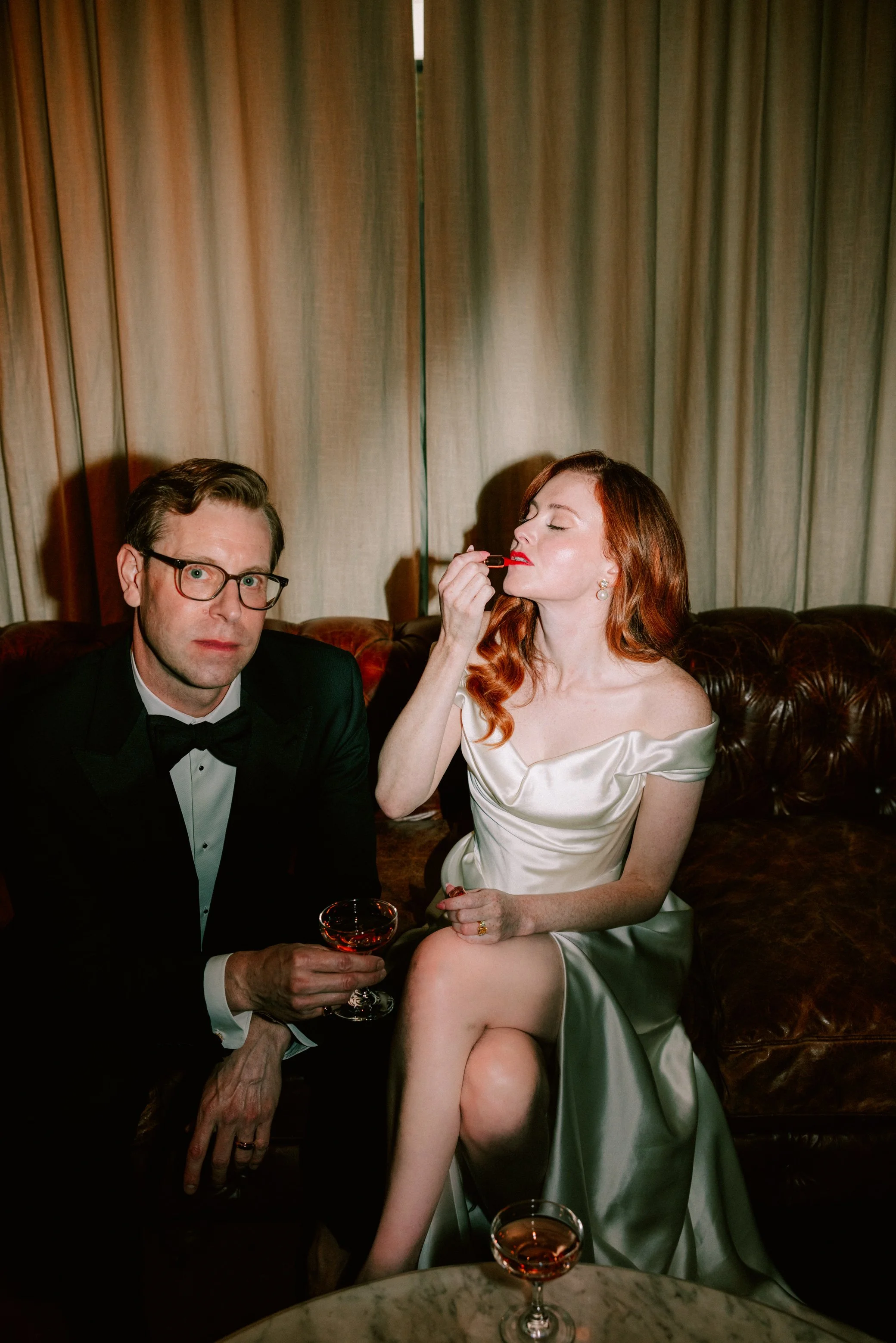 bride puts on red lipstick while groom holds a cocktail coupe glass sitting on leather tufted couch in the lounge of senate garage