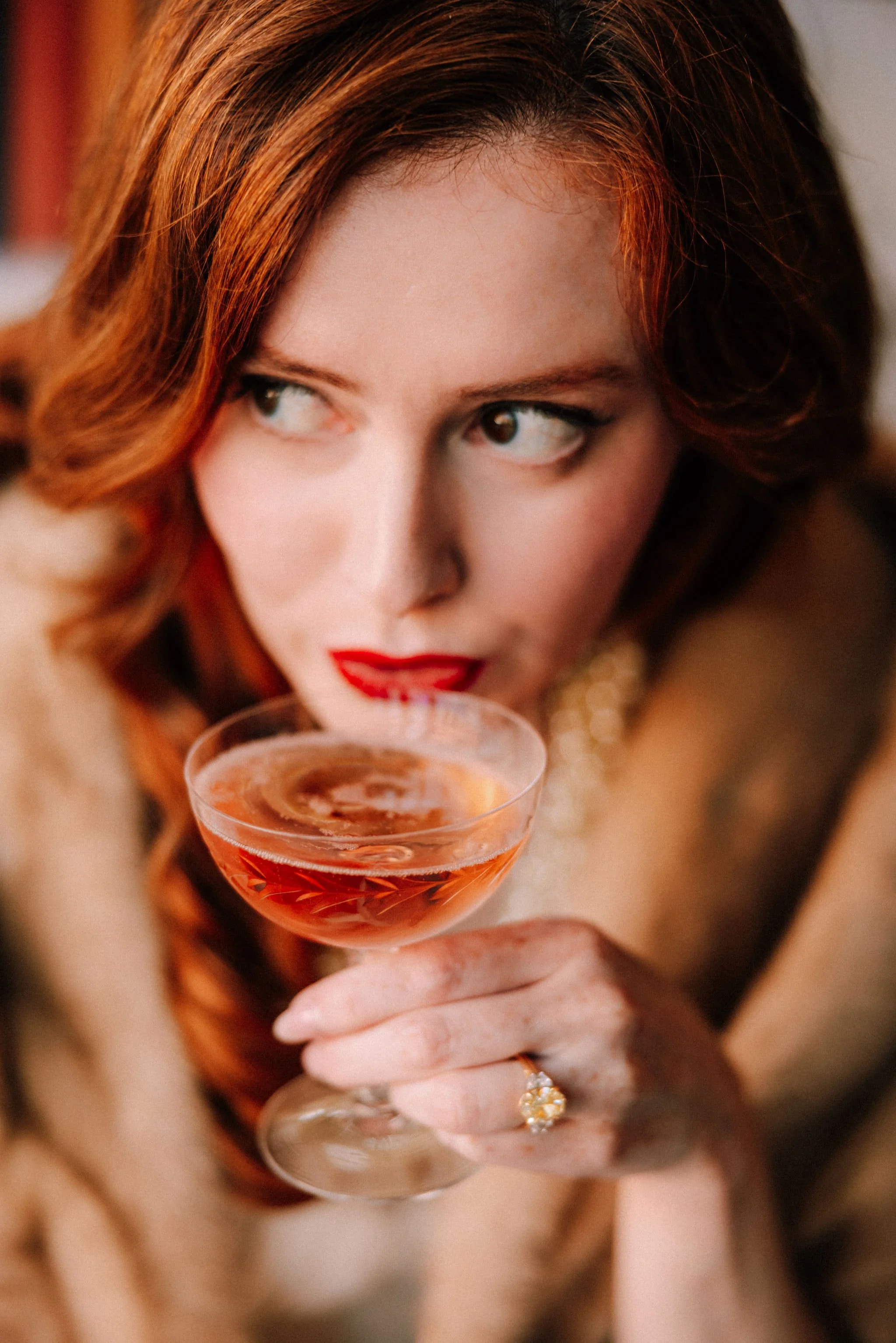 woman with red hair wearing an yellow diamond engagement ring holds a coupe glass with rose sparkling wine up her to lips in brunette wine bar in kingston new york during her engagement photography shoot