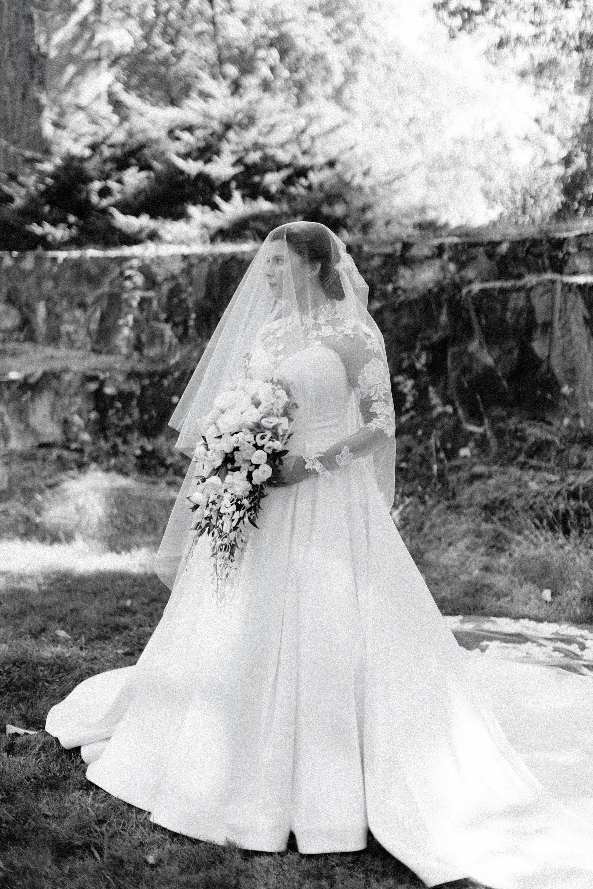 Black and white photo of a bride in a wedding dress holding a bouquet of flowers, standing outdoors with trees and a wooden fence in the background on her wedding day in the hudson valley.