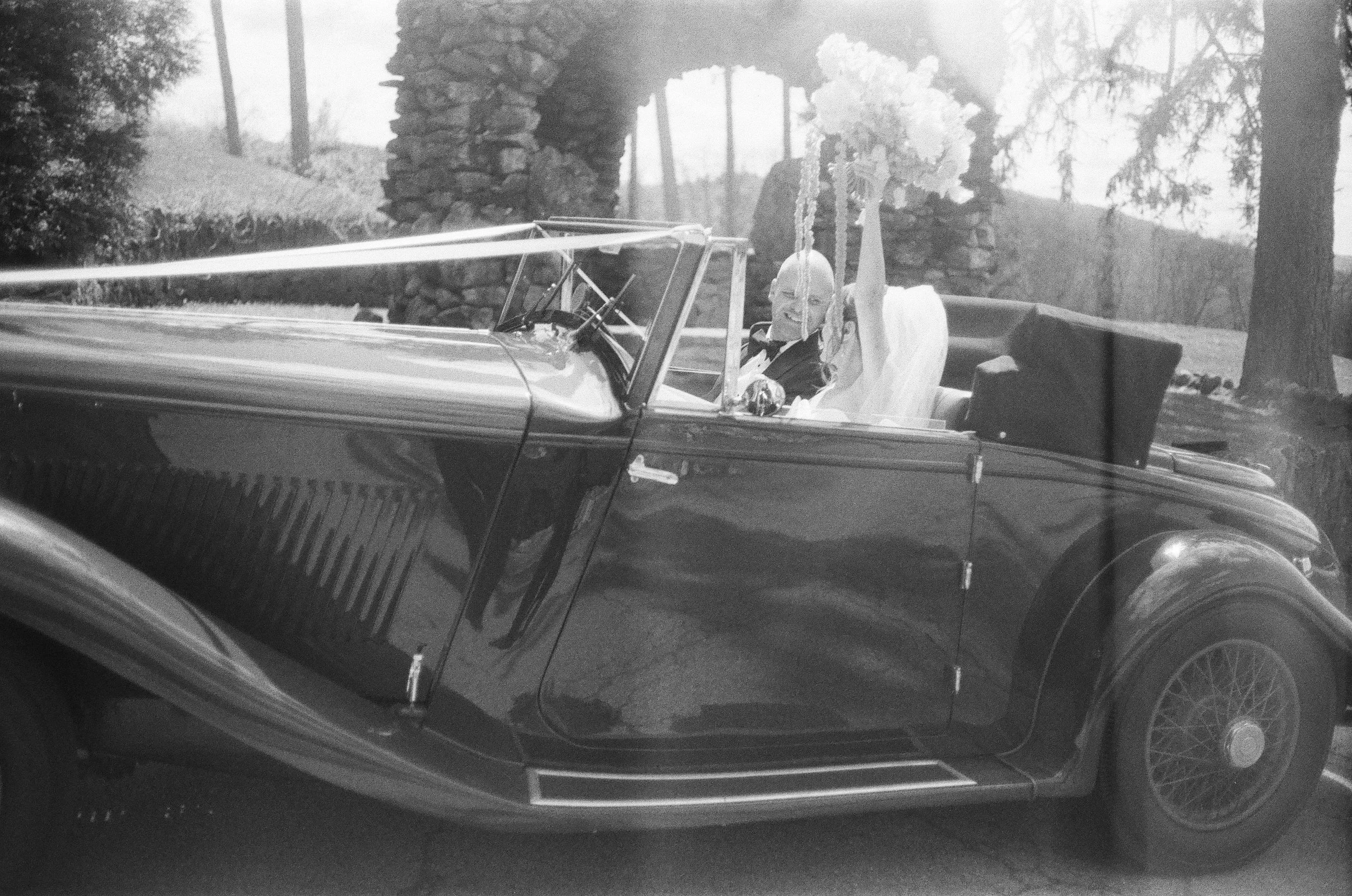 film photograph of bride and groom in vintage car while they look at each other and she holds her bouquet above her head on their wedding day in garrison new york