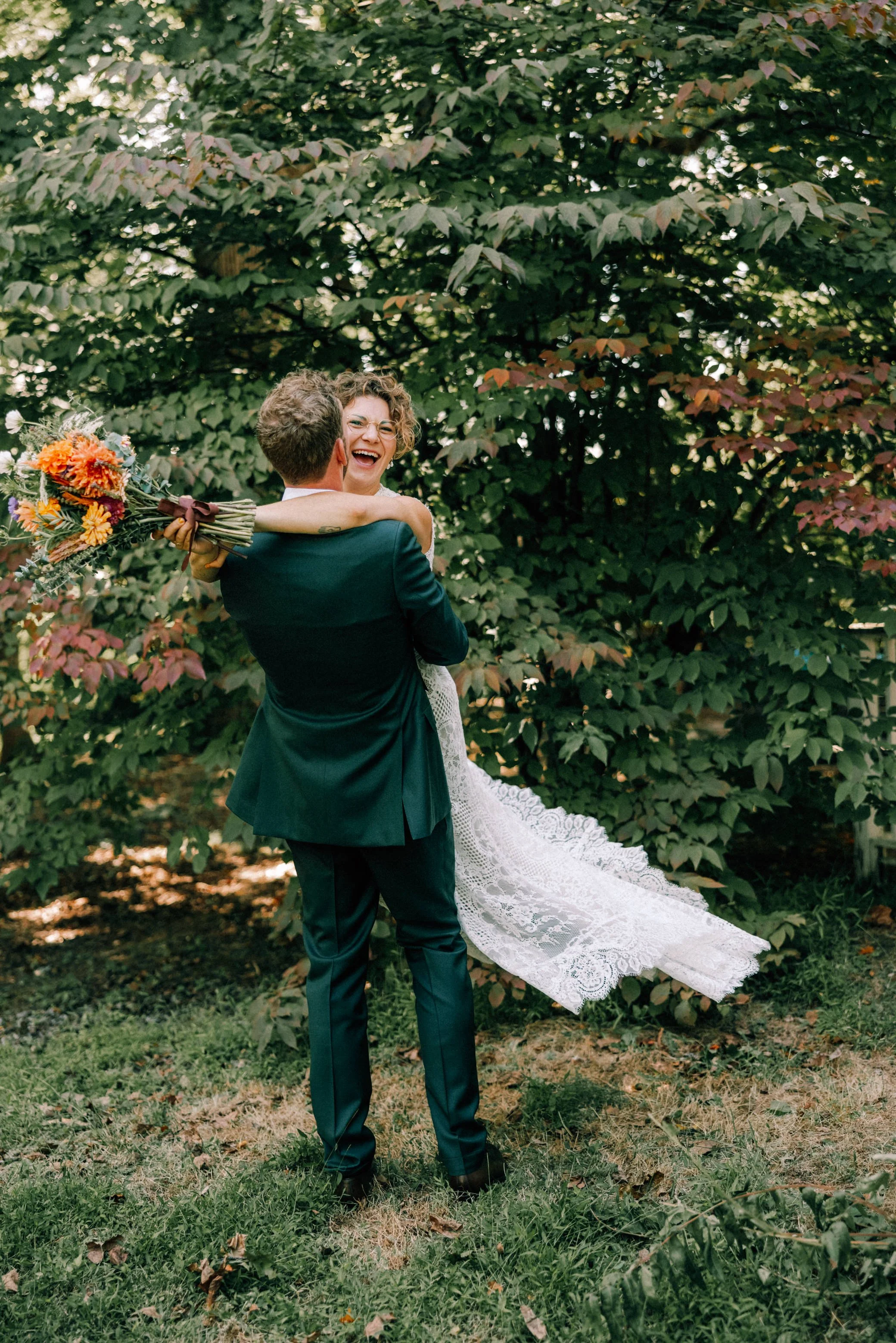 photograph of groom wearing green suit picking up and spinning his bride wearing a lace dress and holding a bouquet while she laughs and smiles at the camera in beacon ny
