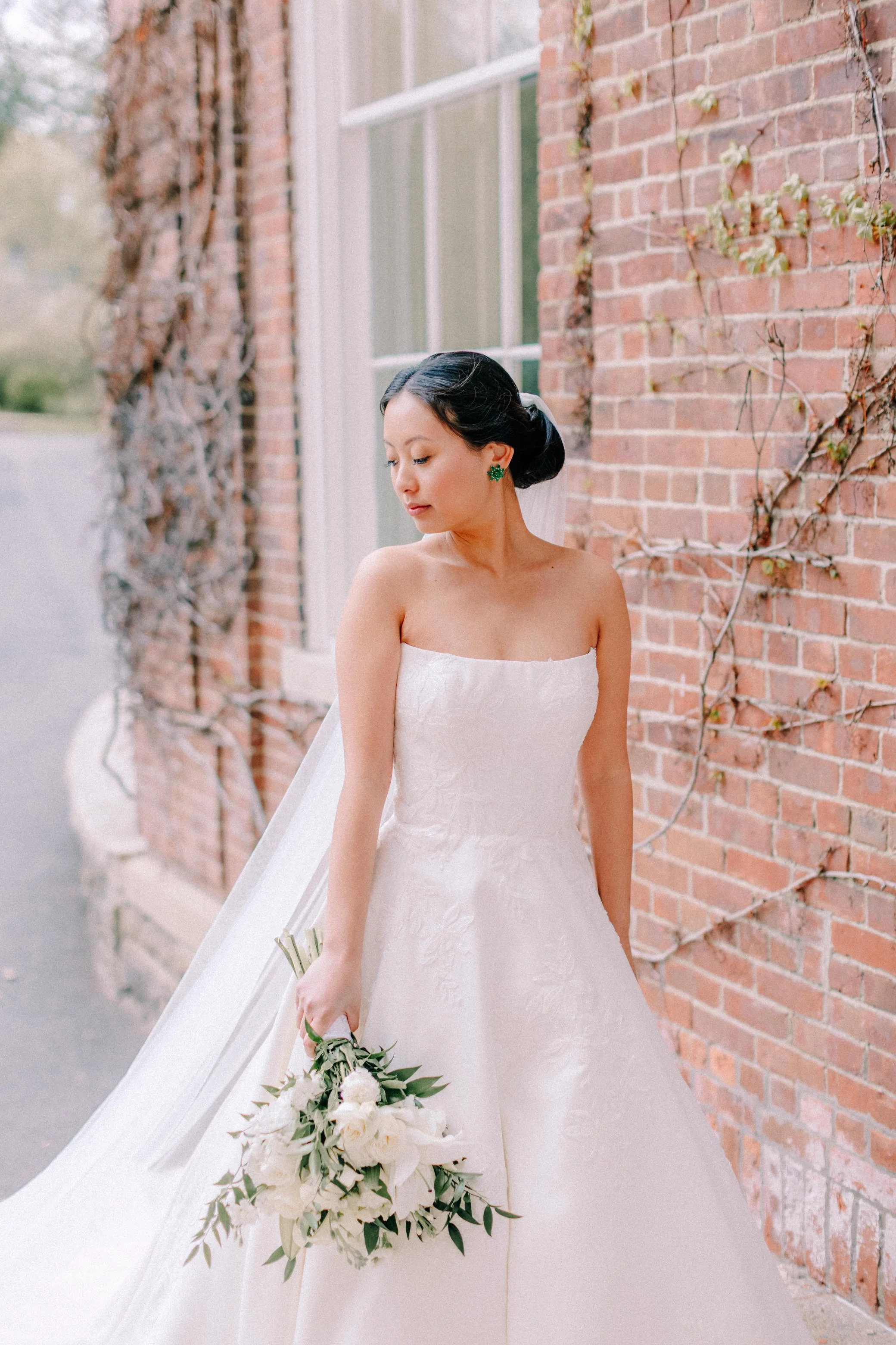 bride looking down towards the right and holding her bouquet in front of a window and brick exterior with vines 