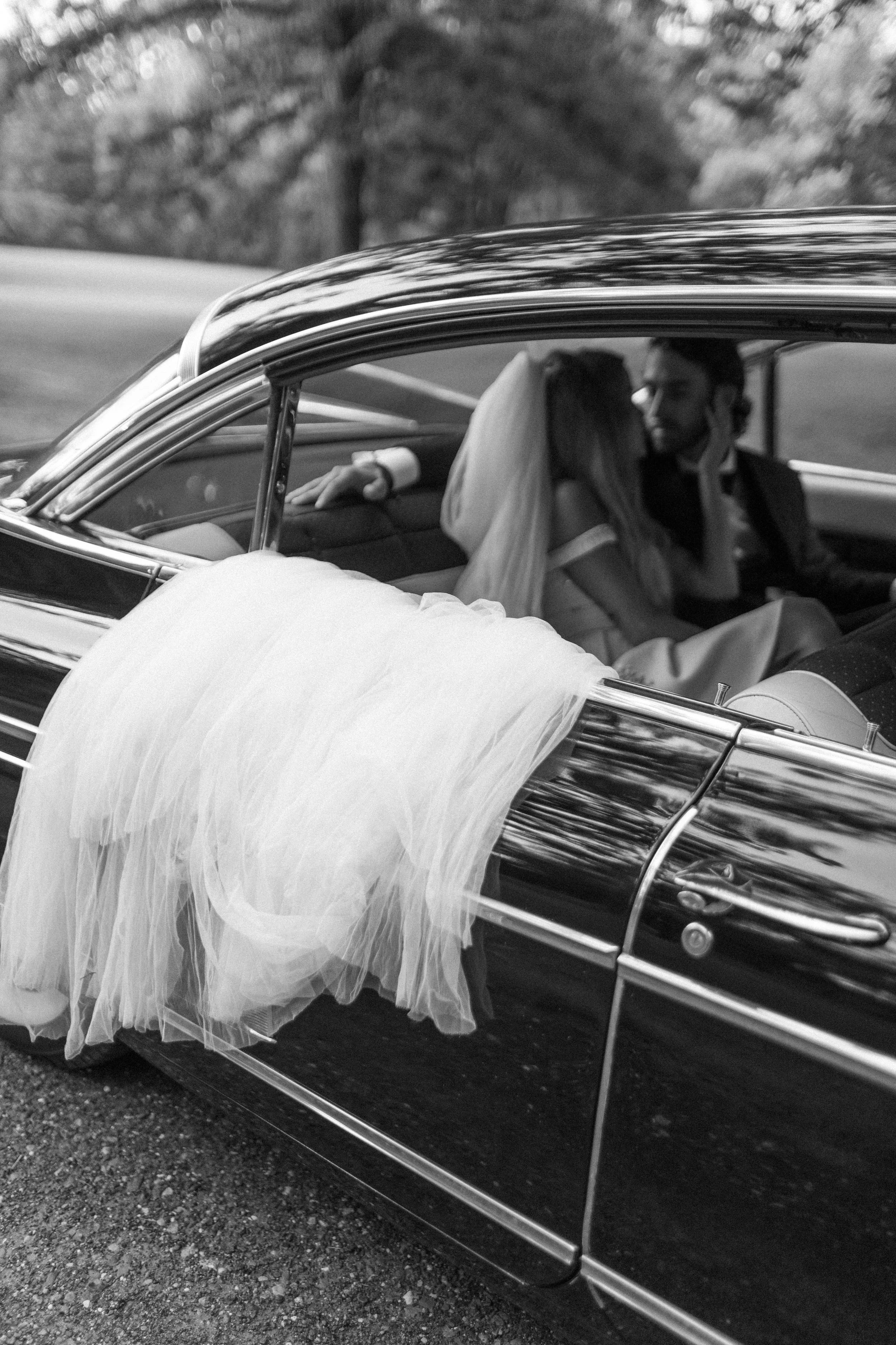 couple almost kiss in the back seat of a black vintage cadillac while the bride's veil hangs out of the door on their wedding day in the hudson valley