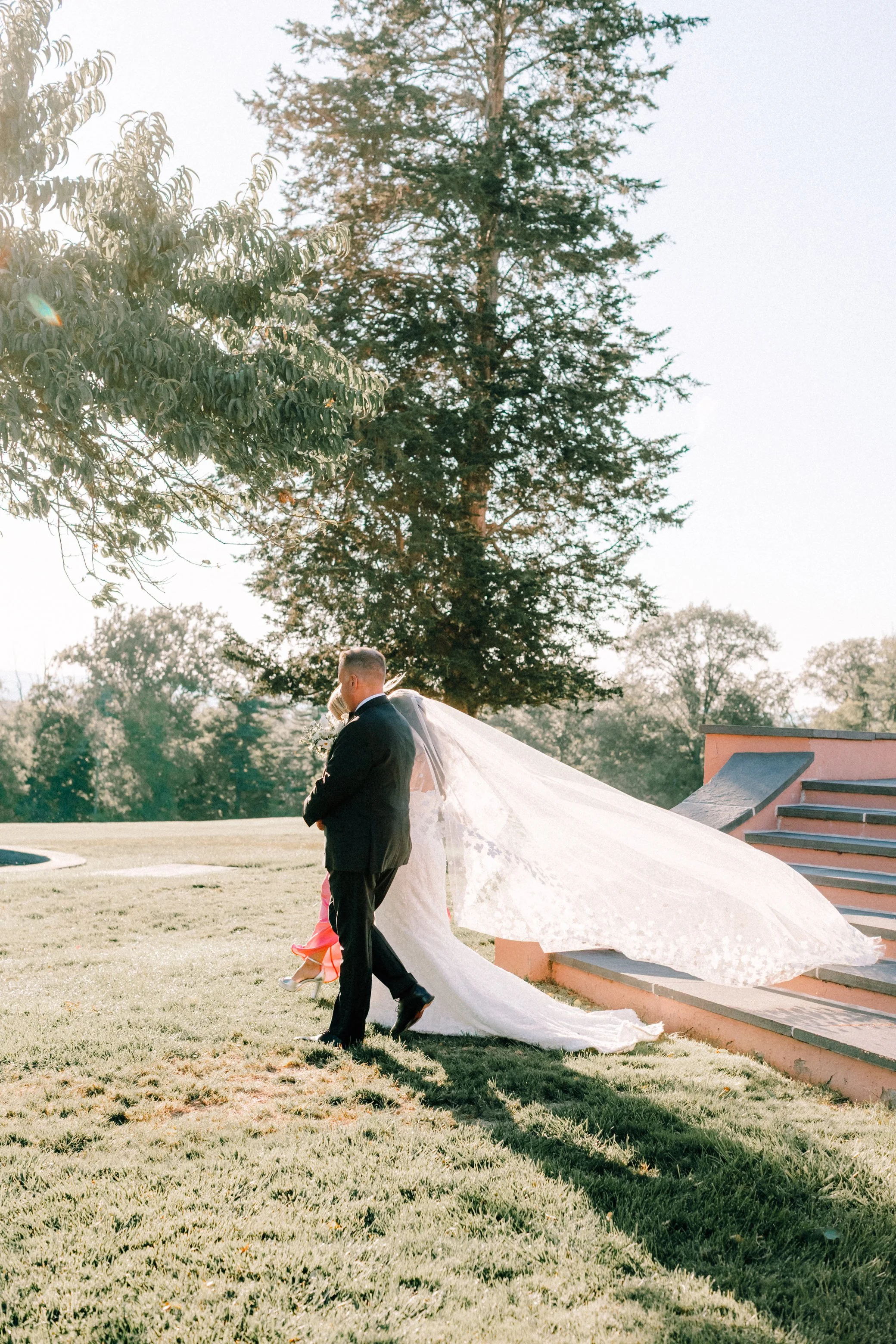 photograph of bride being walked down the aisle by her parents while her cathedral length veil illuminated by the sun is floating in the air on her wedding day at glenmere mansion
