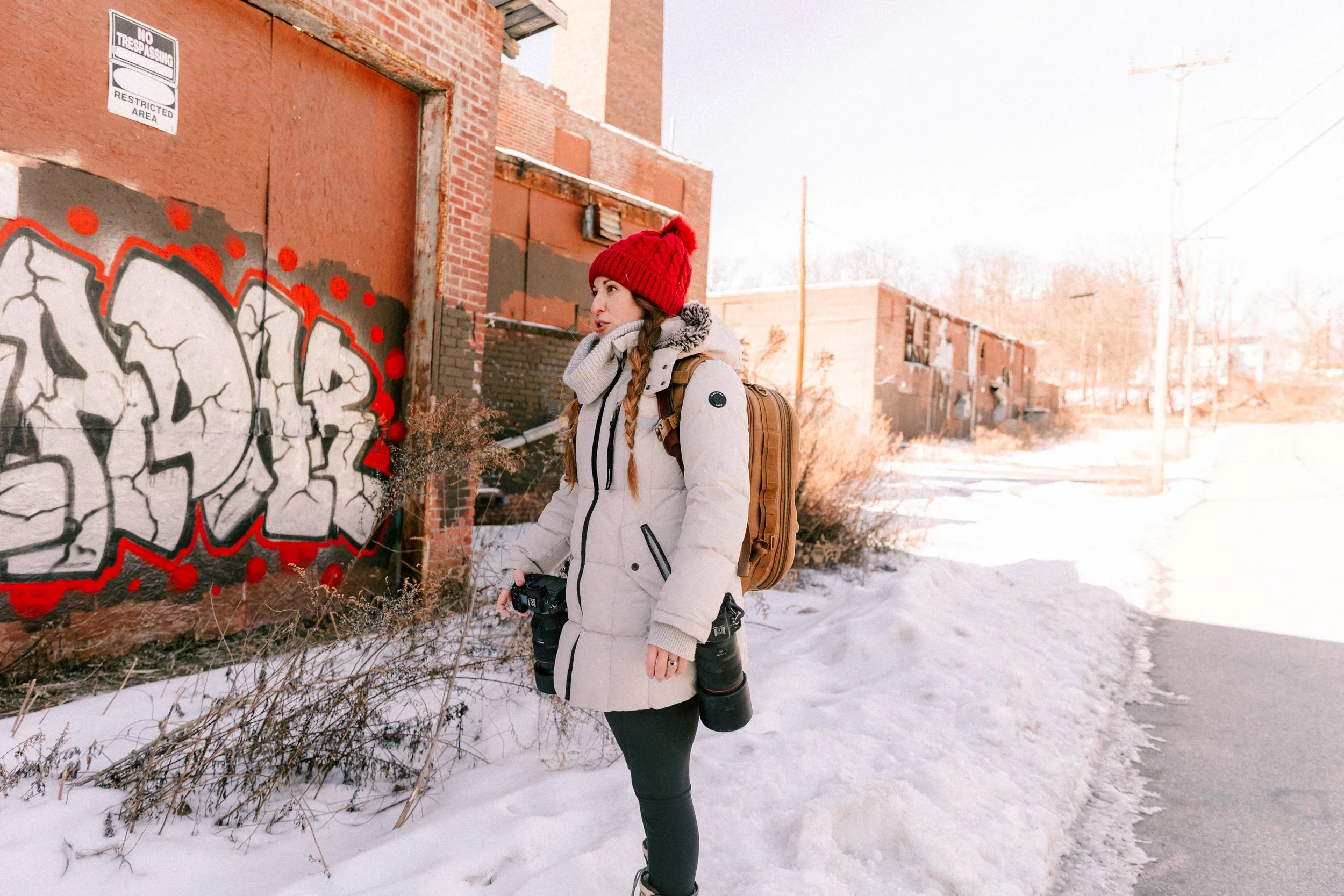 Caitlinn Mahar-Daniels, hudson valley wedding photographer, dressed in winter clothing with a red knit hat and a backpack, standing outdoors in a snow-covered area at Madam Brett Park in Beacon, NY. She is holding a camera with a large lens.