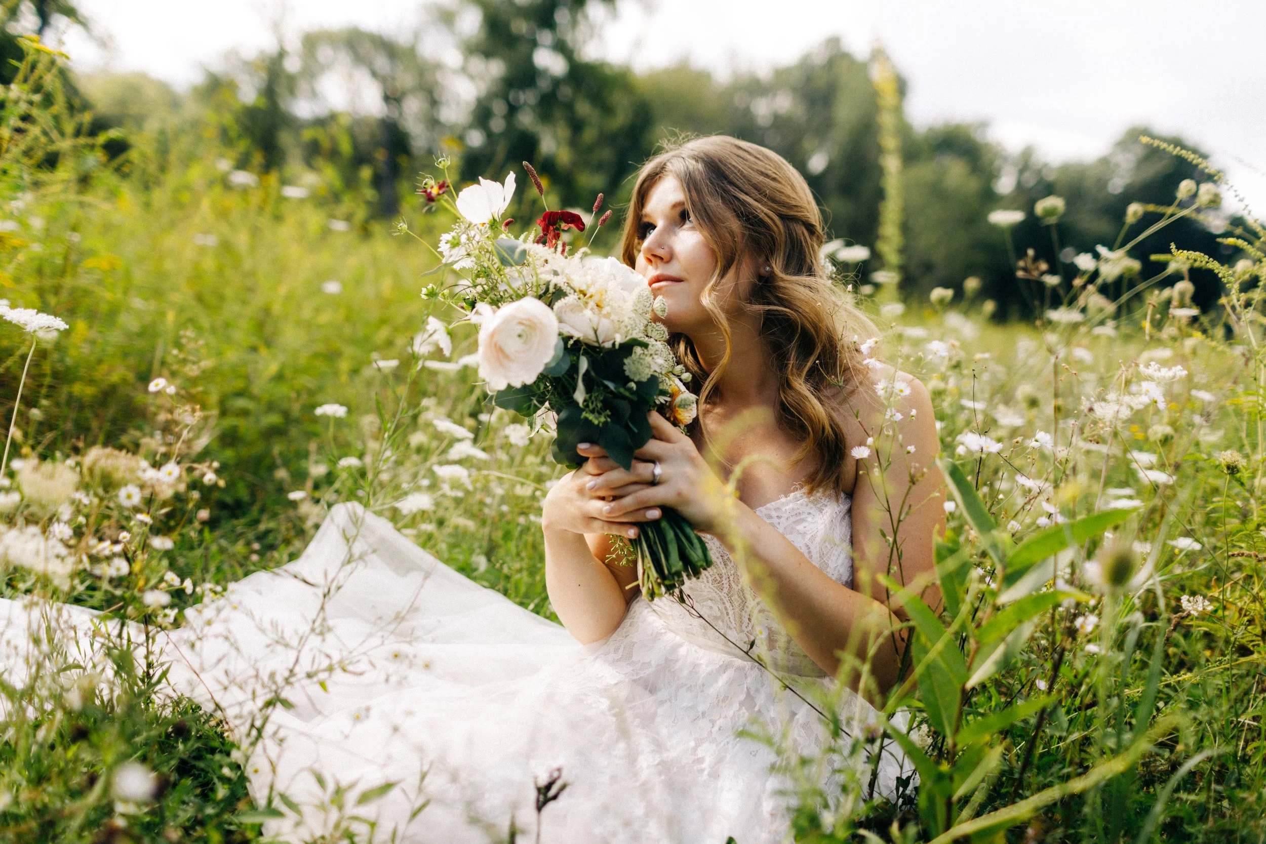 bride sits in wild flowers while holder her bouquet up to her face and looking into the distance on her wedding day at blooming hill farm
