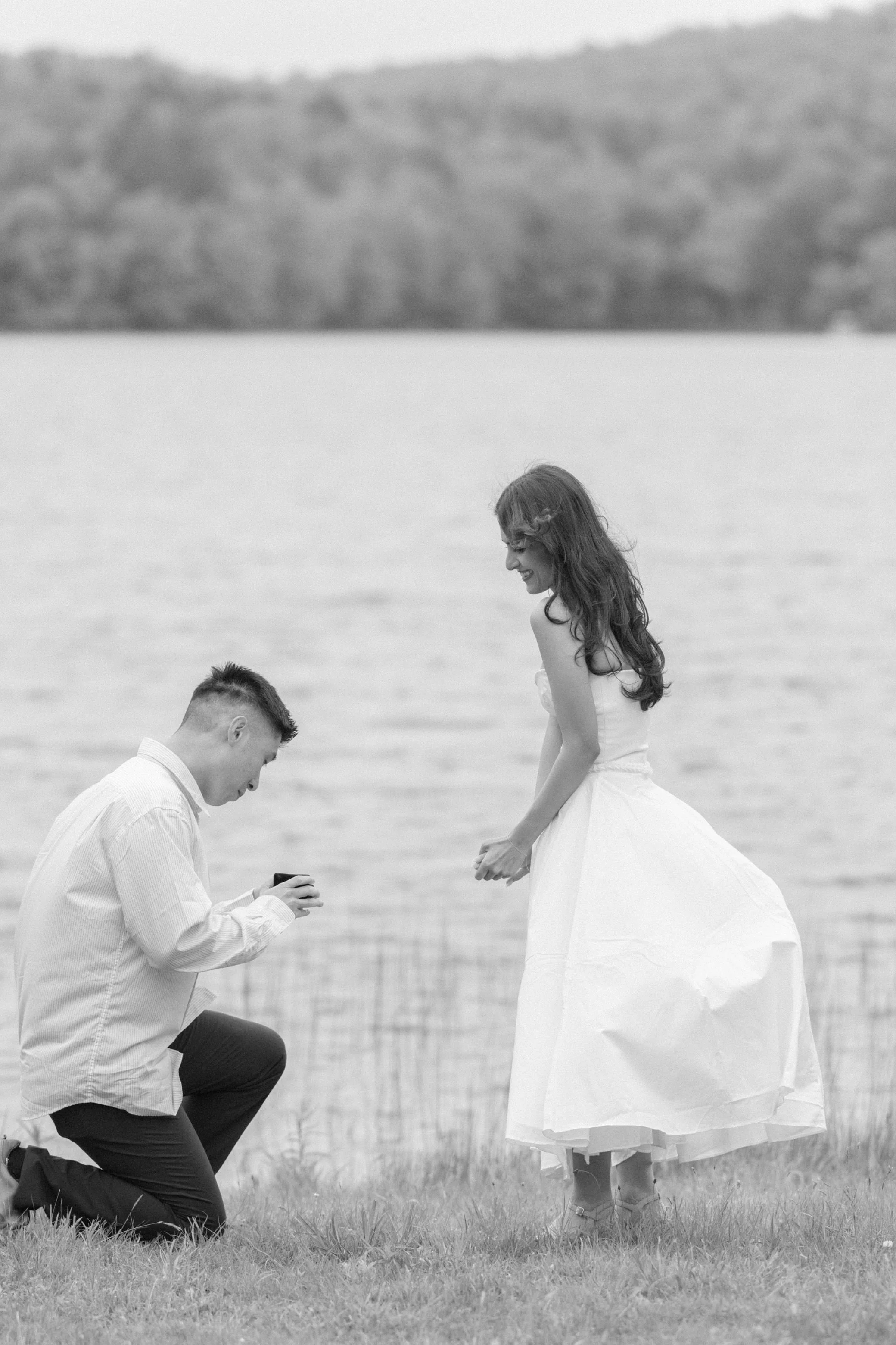black and white photo of man down on one knee proposing to woman in a white dress with kenoza lake behind them
