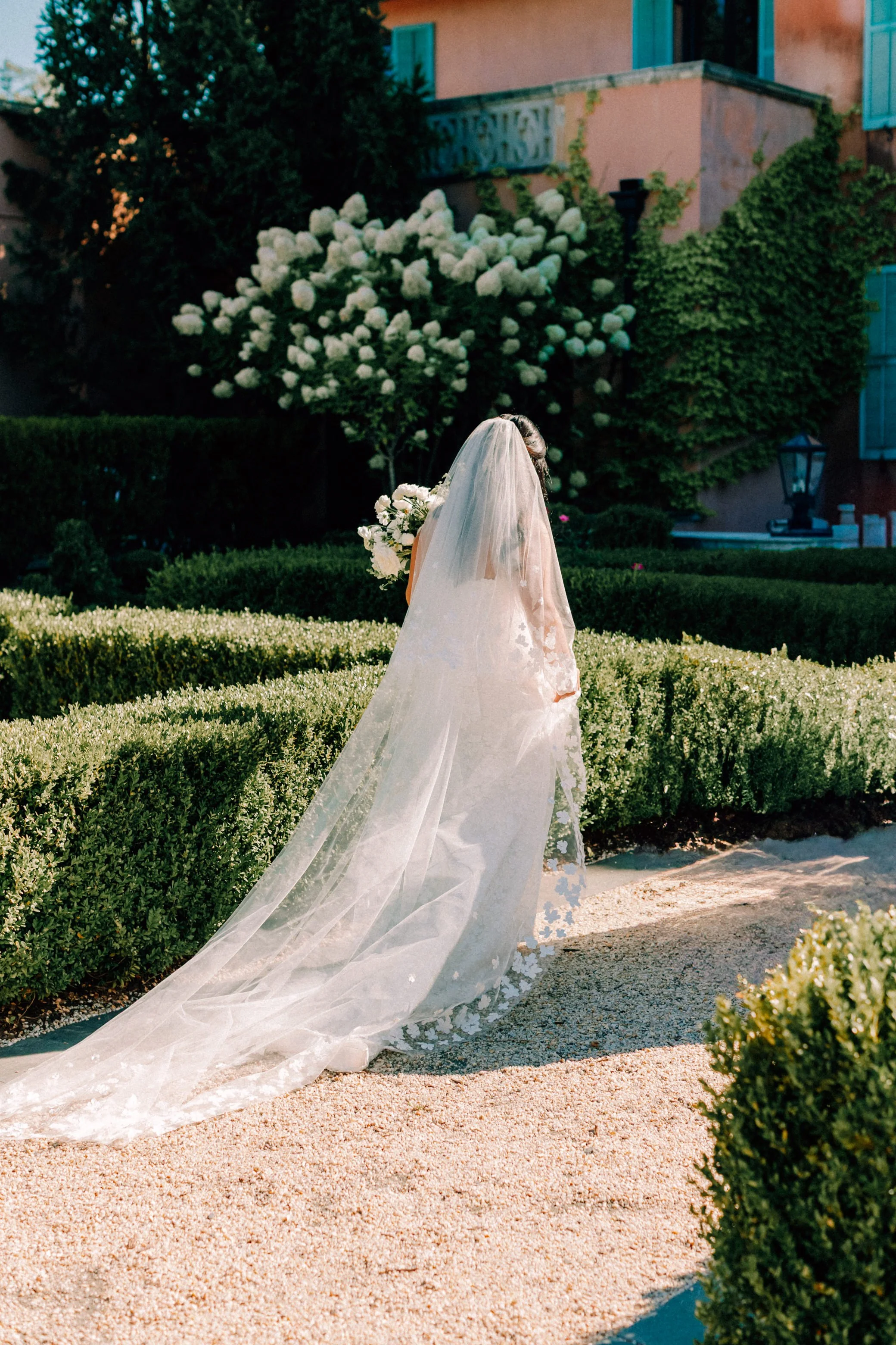 bride with floral veil walking through formal gardens while holding her bouquet at glenmere mansion on her wedding day