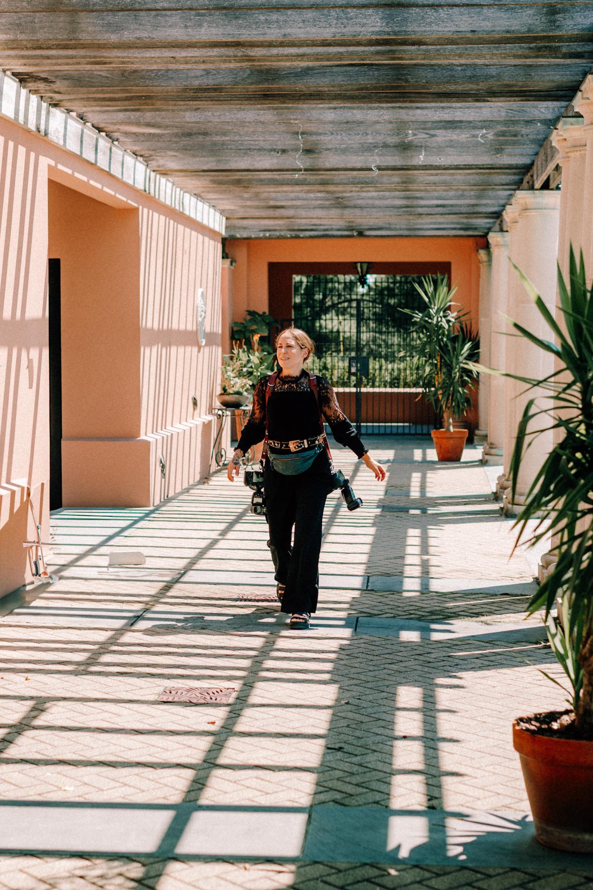 Caitlinn Mahar-Daniels, hudson valley wedding photographer, walking in a sunny outdoor corridor with potted plants, shadows from a pergola, and peach-colored walls at Glenmere Mansion.