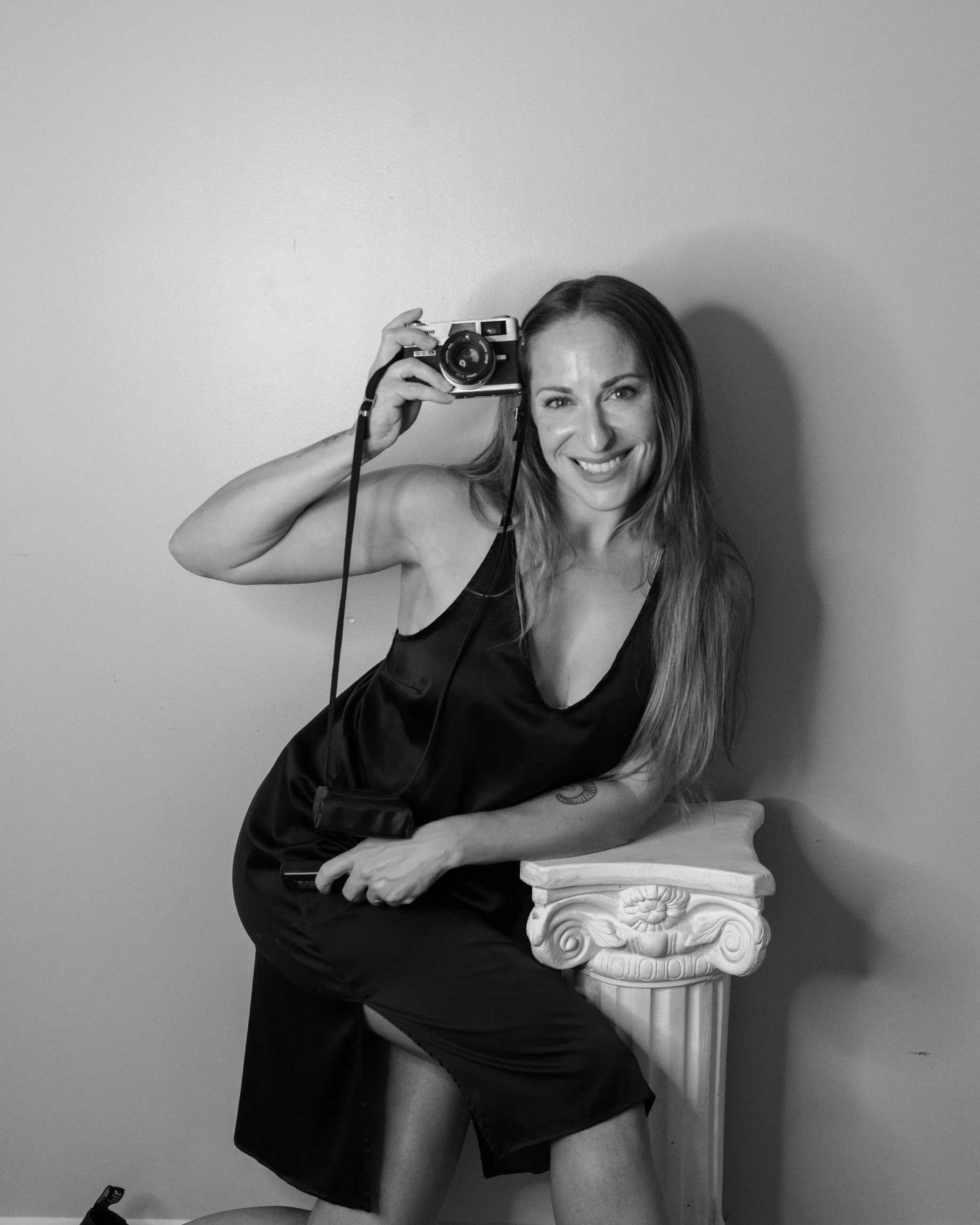 Black and white photo of Caitlinn Mahar-Daniels with long hair, wearing a sleeveless dress, sitting on a stool with her arm resting on a decorative pillar, holding a vintage camera up to her eye, looking at the camera.