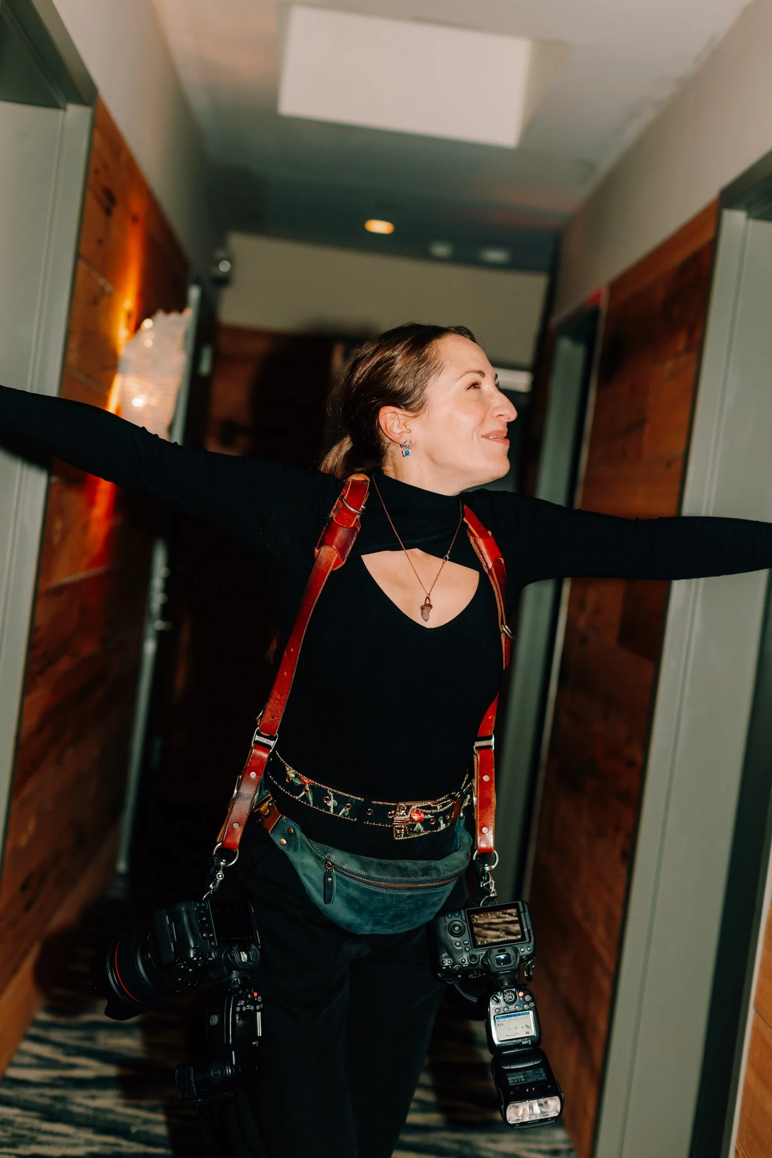 Caitlinn Mahar-Daniels, hudson valley wedding photographer, with arms outstretched, smiling, in a hallway with wooden wall paneling and ceiling lights at The Roundhouse.
