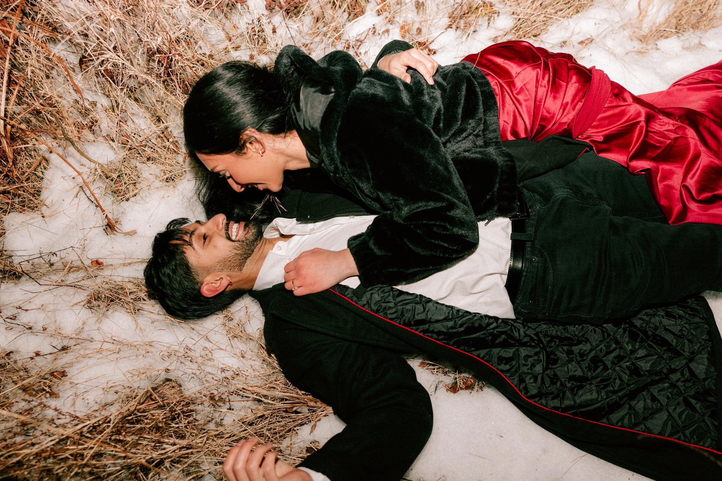 A man and woman lying on the snow and dry grass, smiling and looking at each other during their adventurous engagement photography shoot at Minnewaska State Park.