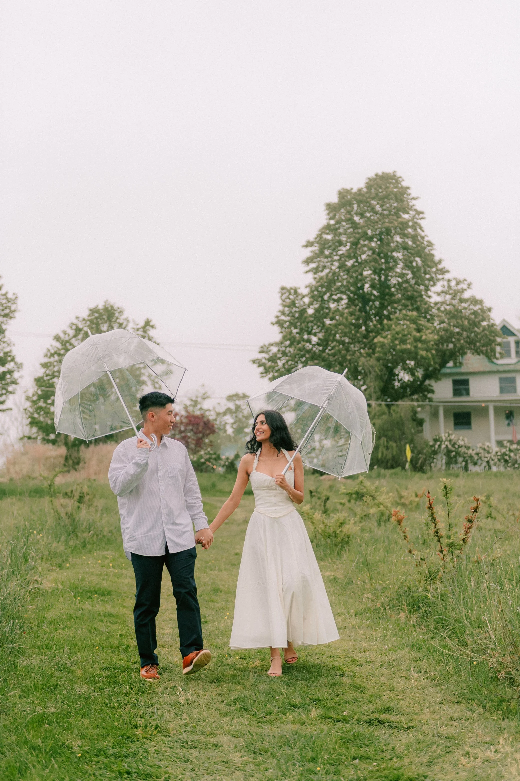 newly engaged couple hold hands and walk down a grassy hill in the rain while holding clear umbrellas at kenoza hall