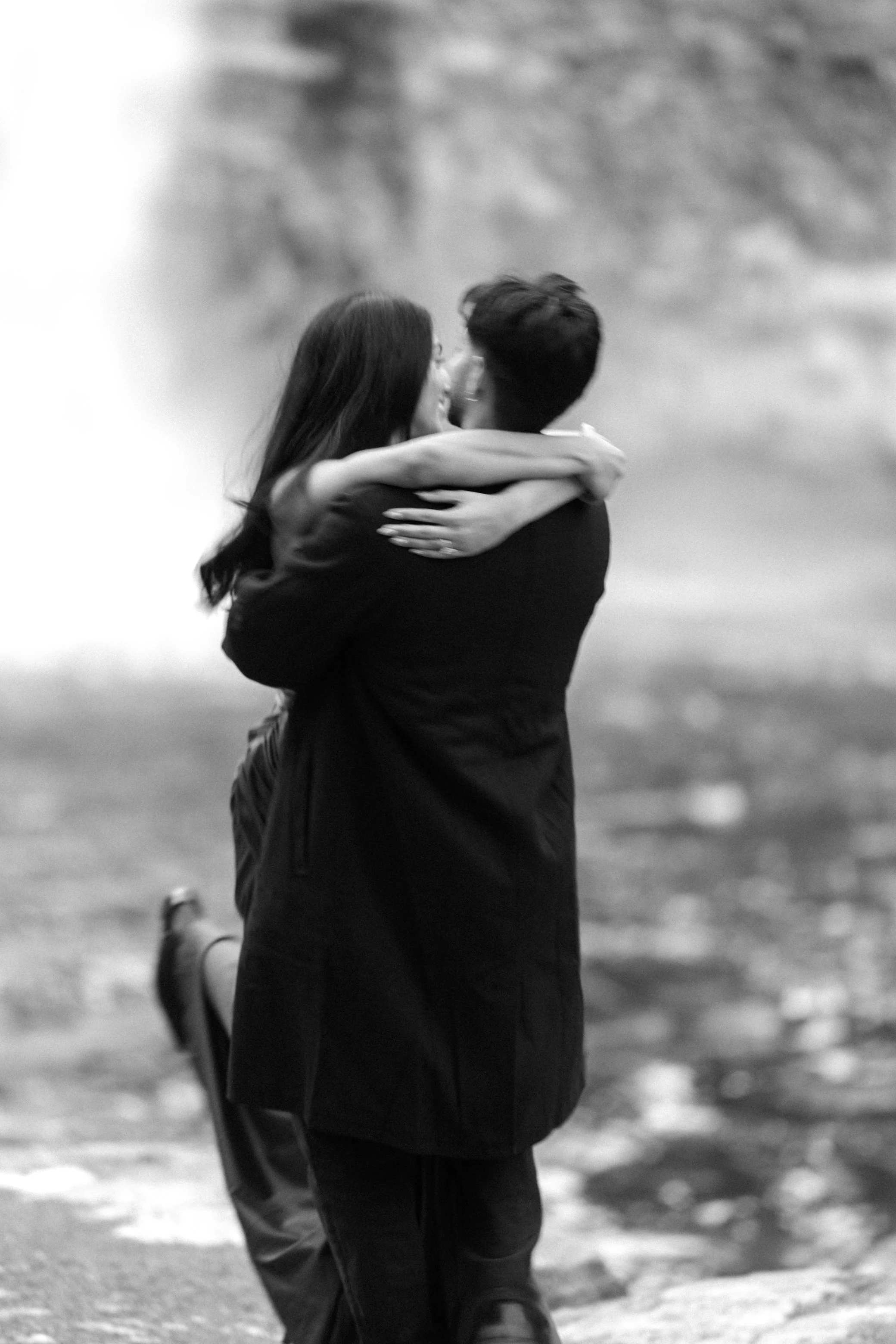 A black and white photo of a couple embracing, with the woman being lifted off the ground by the man, during their romantic winter engagement photo shoot at Awosting Falls in Minnewaska State Park.