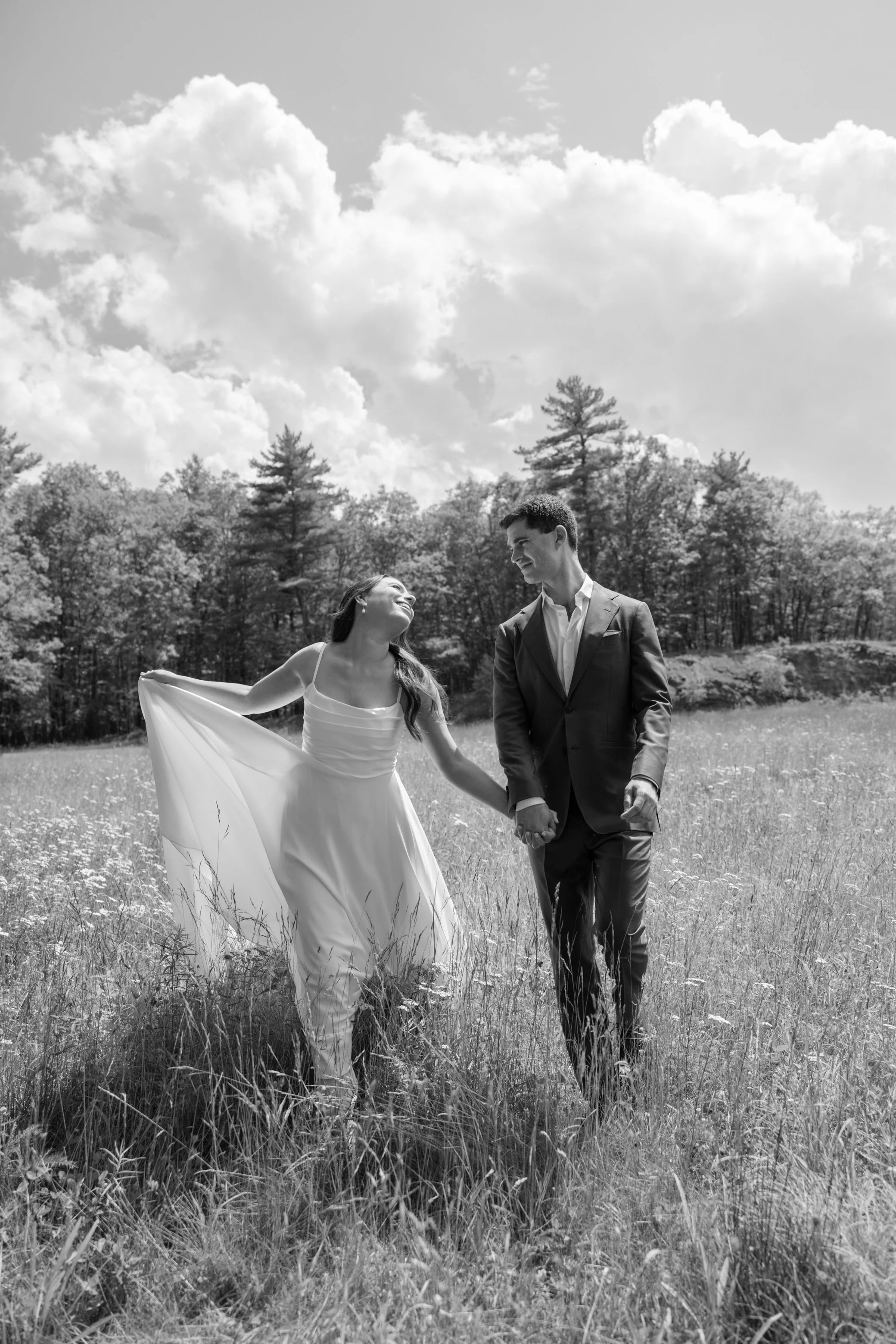 couple walk through field of wildflowers while bride holds her dress while laughing on their wedding day at gather greene