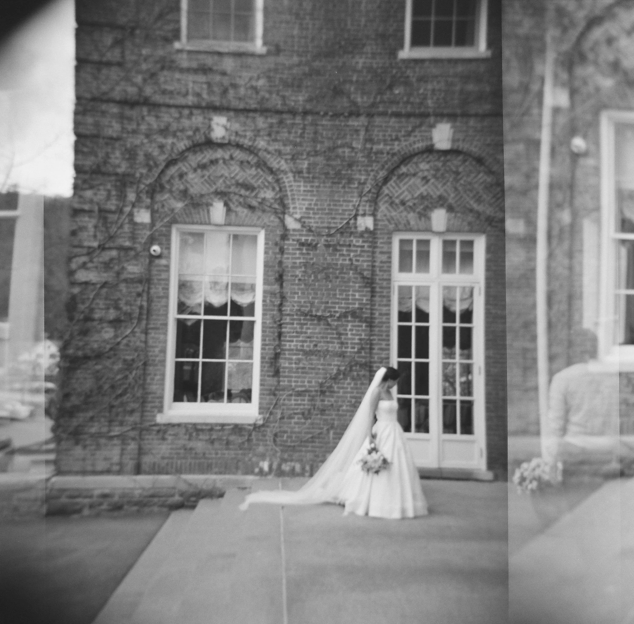 film photograph with light leaks and double exposure of bride standing in front of brick building with big windows on her wedding day at otesaga hotel