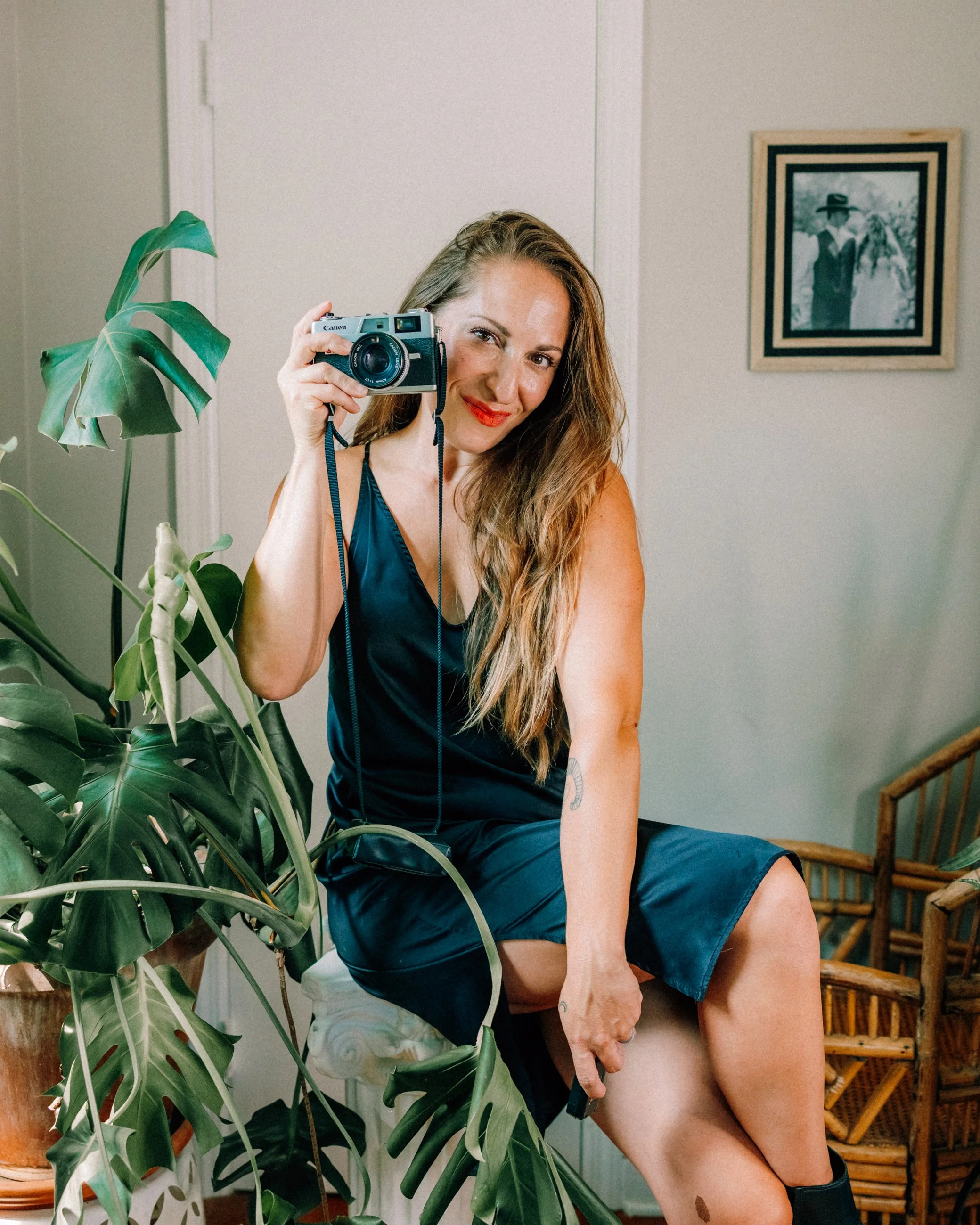 Hudson Valley Wedding Photographer Caitlinn Mahar-Daniels wearing a black satin dress, sitting on a decorative stool, holding a vintage film camera to her face, smiling, surrounded by green indoor plants and a wicker chair.