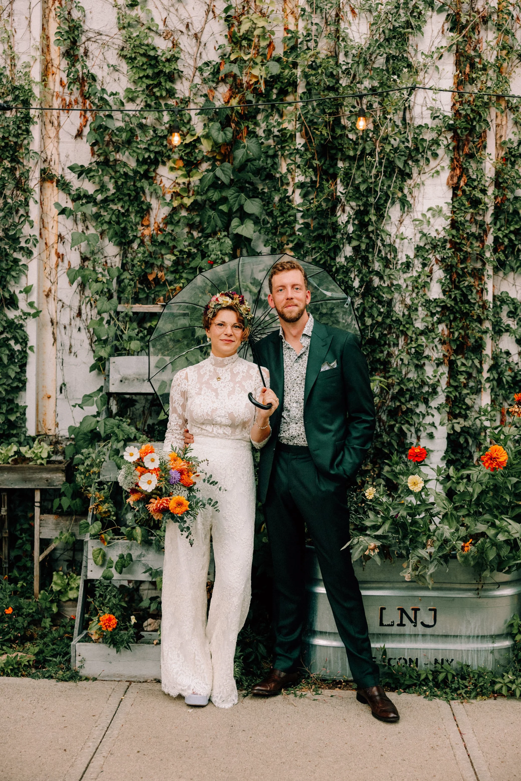 Bride wearing jumpsuit with flower crown holding a bouquet and groom in green suit holding an umbrella on their wedding day at The Yard in Beacon NY