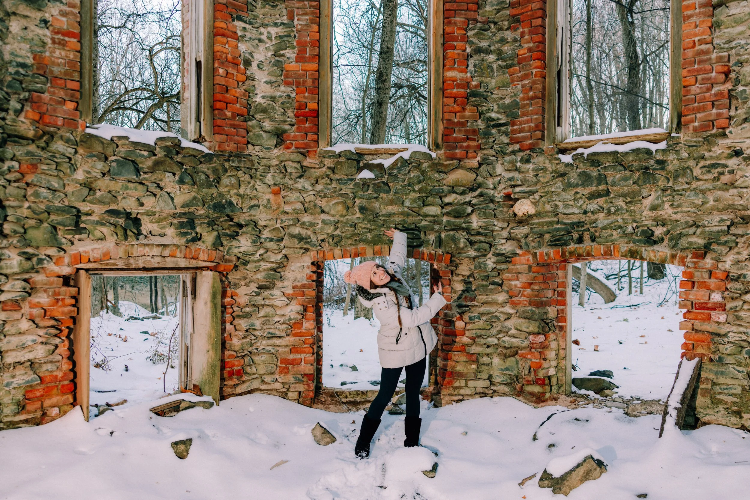 Caitlinn Mahar-Daniels, hudson valley wedding photographer, dressed warmly in winter clothes, including a pink knit hat, is standing inside the ruins of an old brick and stone building in a snowy forest.