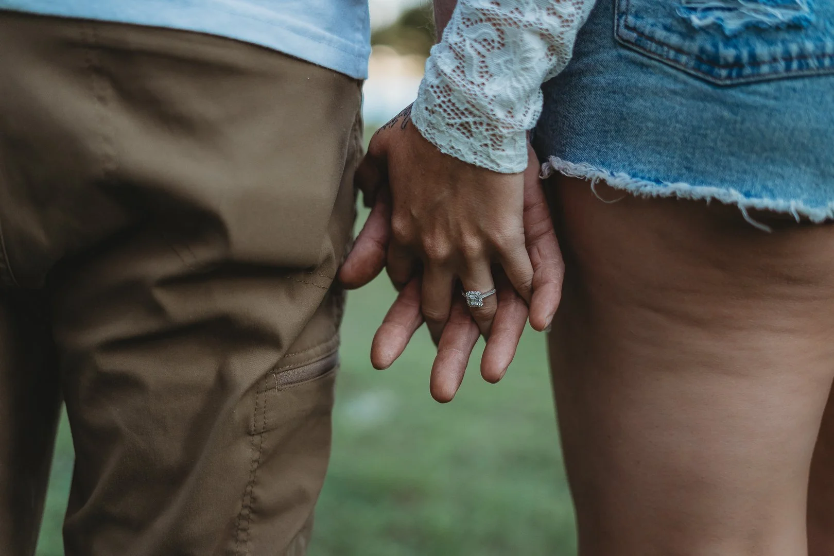 Close-up of a couple holding hands, one wearing an engagement ring, with denim shorts and khaki pants in view.