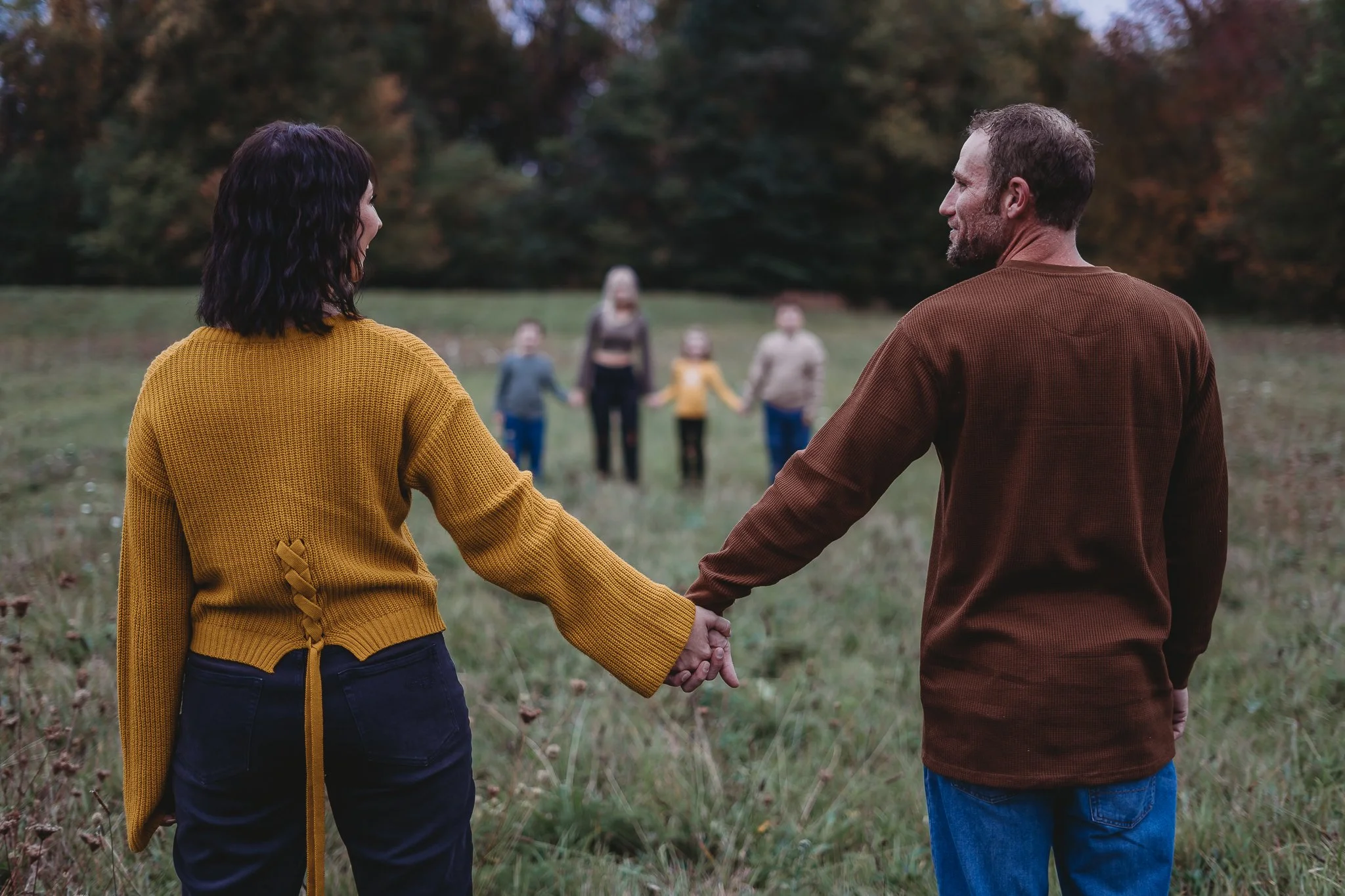 Couple holds hands in a field, facing children in the background.
