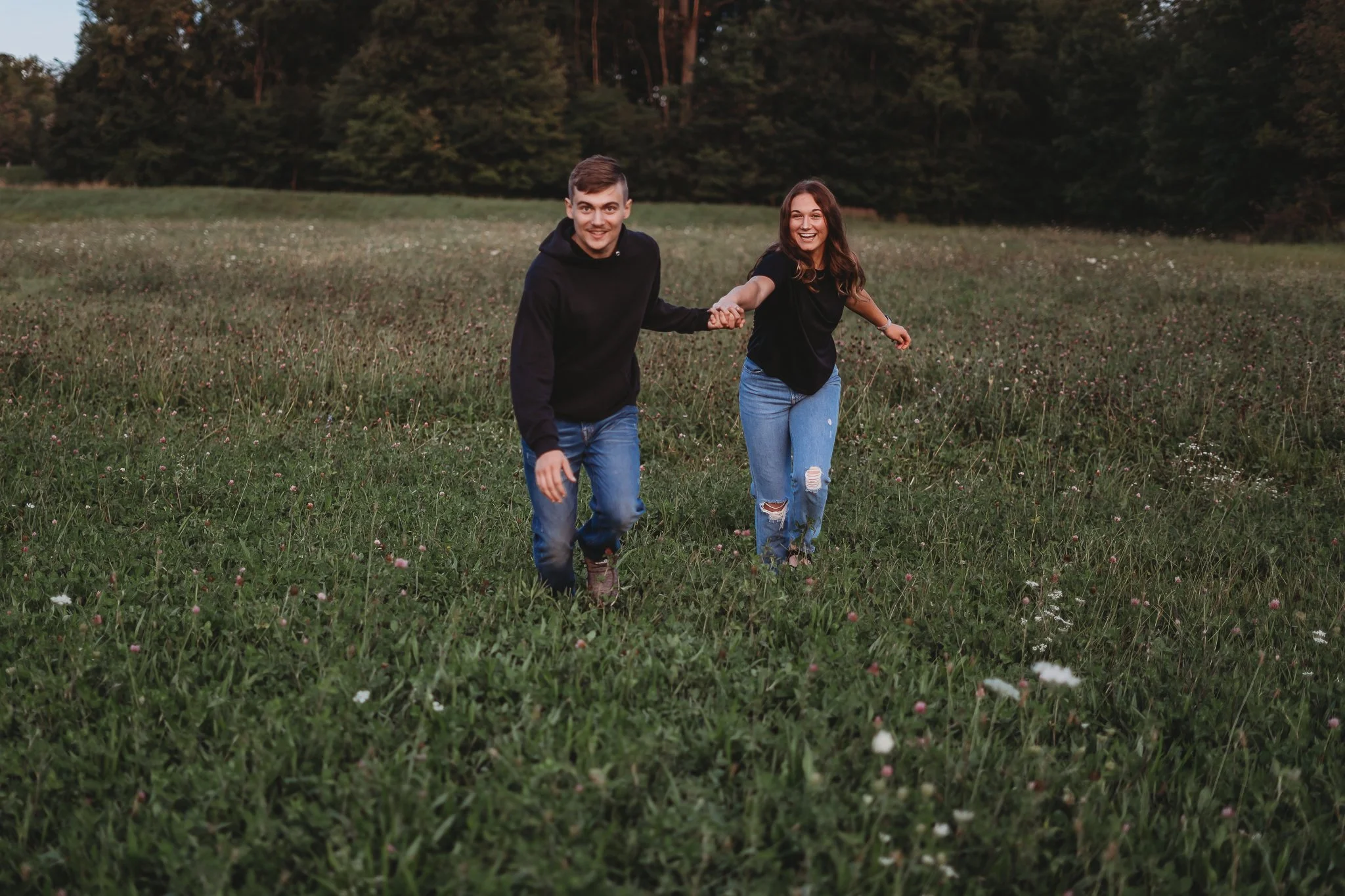 A couple holding hands and running through a grassy field with trees in the background.