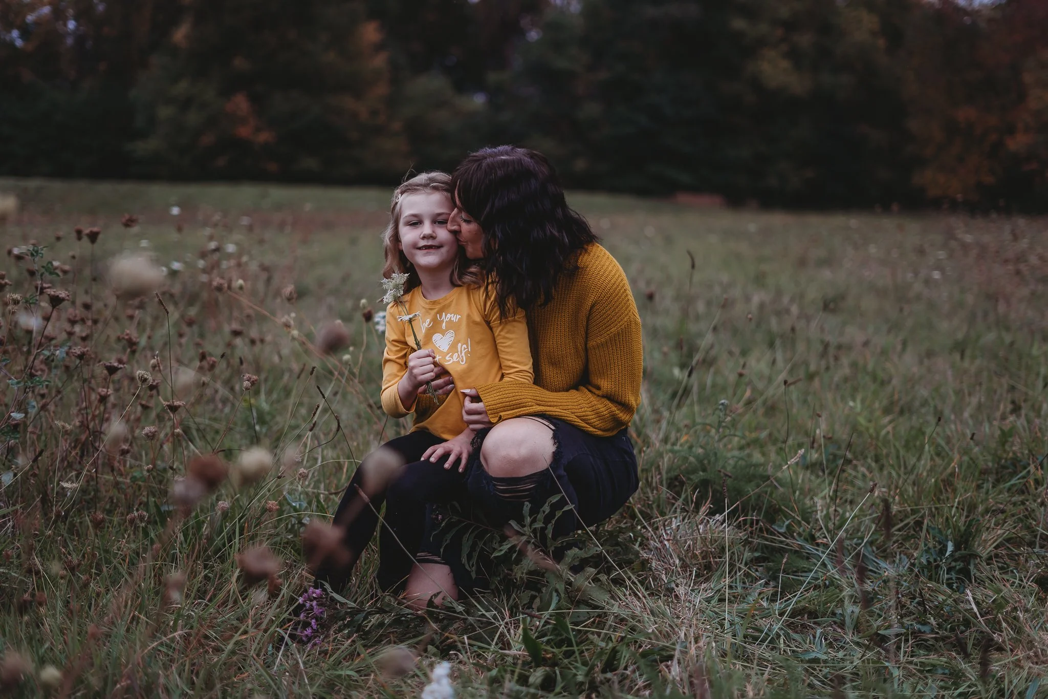 A woman and a child outside in a field, both wearing yellow shirts. The woman is kissing the child's head, and the child is holding a small flower. The setting is a grassy field with blurred trees in the background.