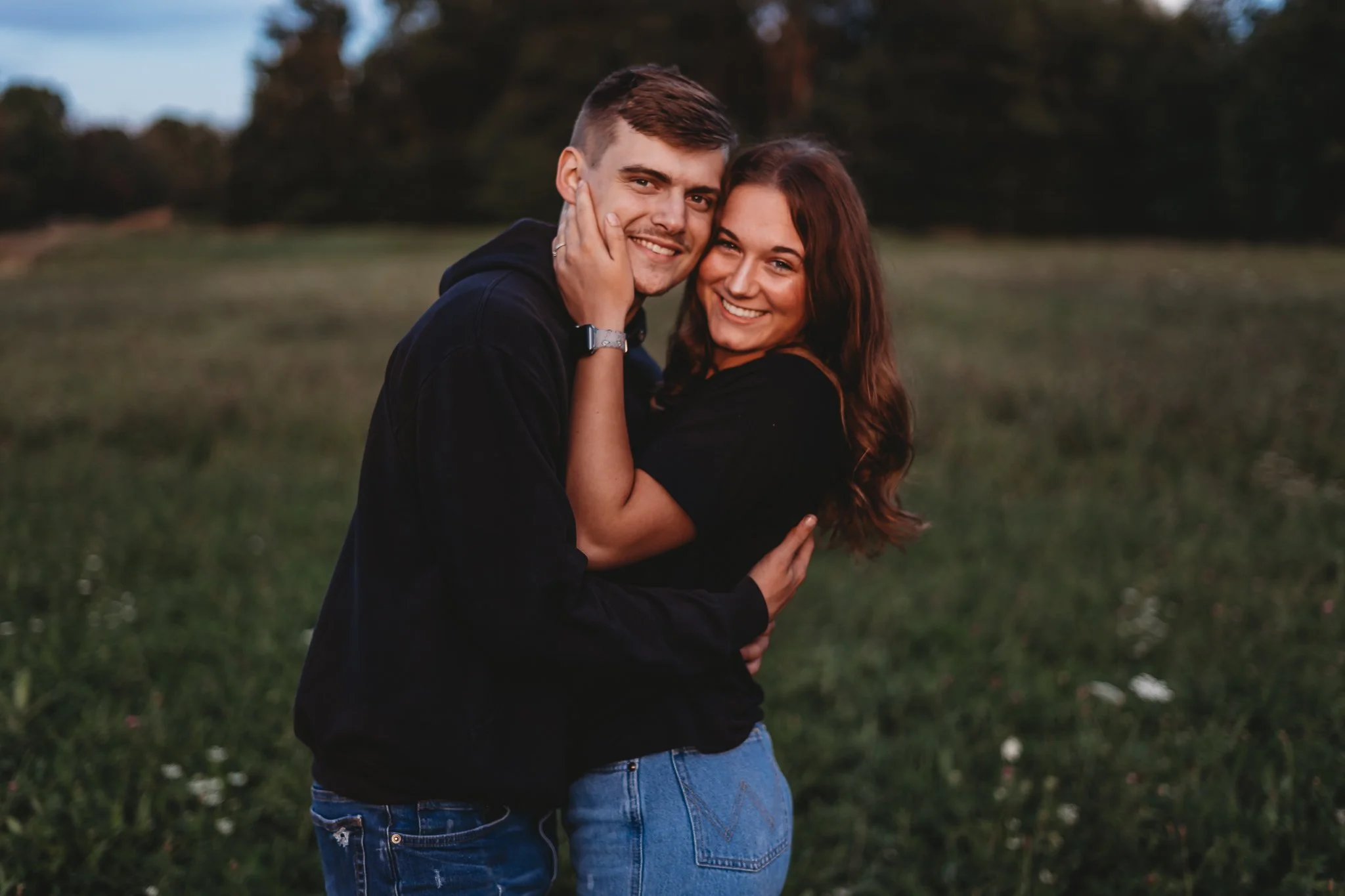 A smiling couple embracing in an open grassy field with trees in the background.