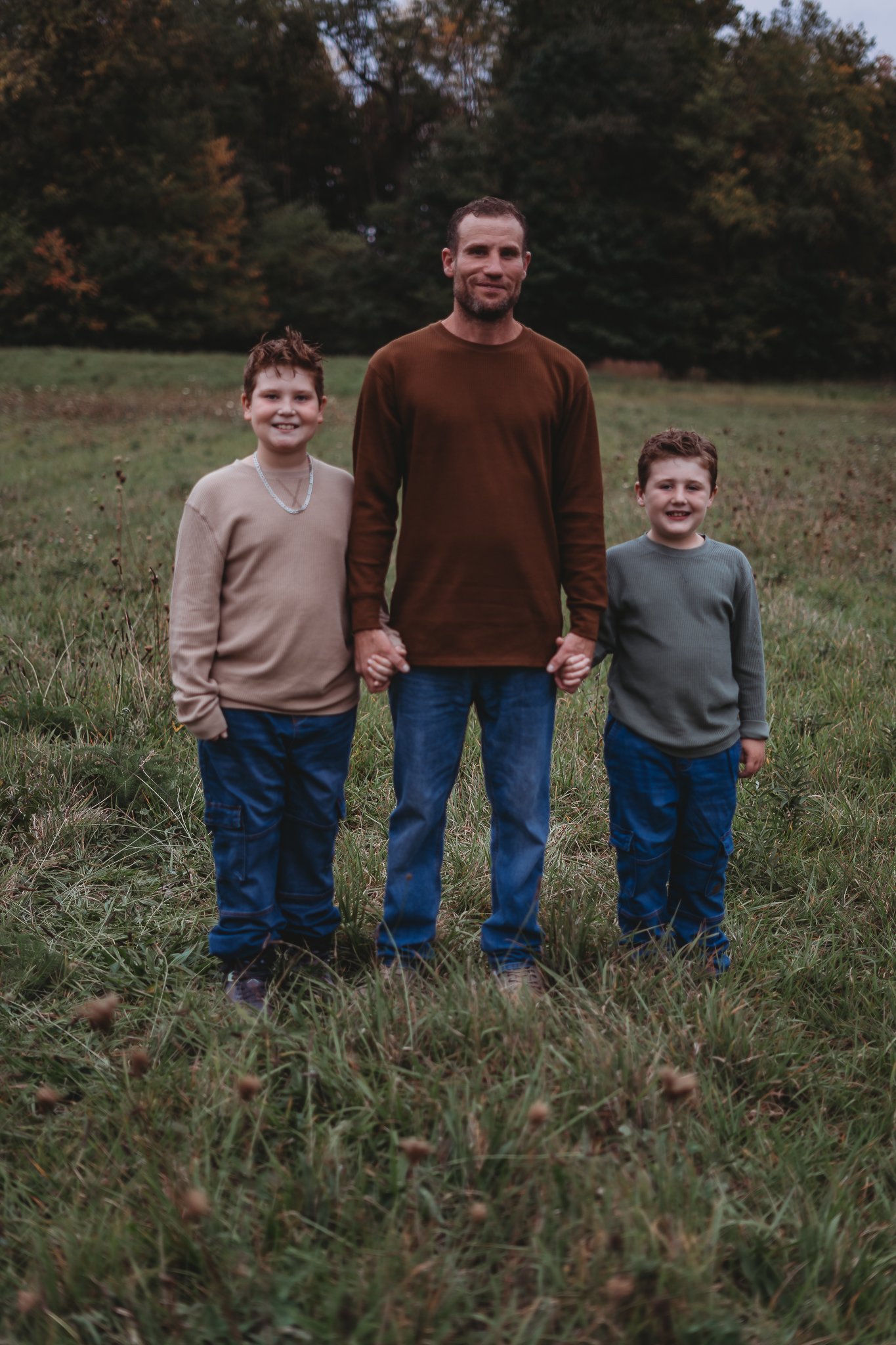 A man standing with two children in a grassy field, holding their hands. Trees are visible in the background.