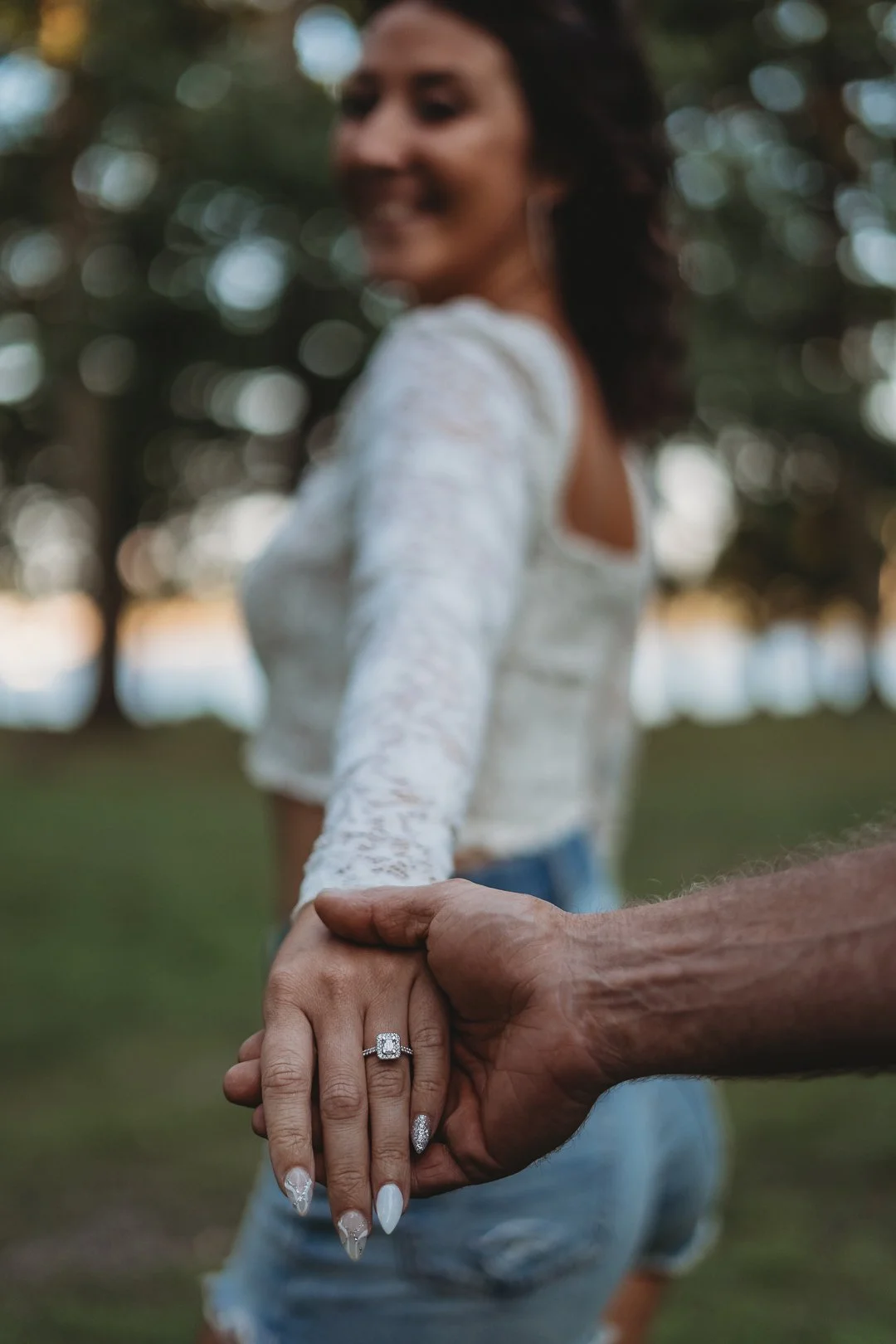 A woman wearing a lace top and denim shorts holding hands with a man, displaying an engagement ring, outdoors.