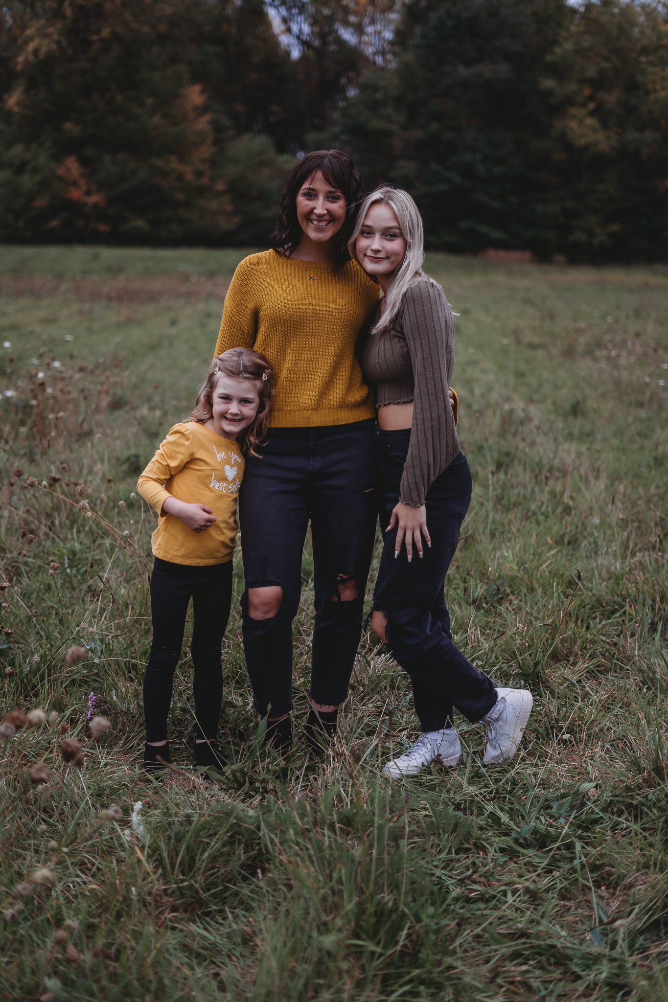 Three people, two adults and one child, standing together on grass in an outdoor setting. The child wears a yellow shirt, an adult in a mustard sweater, and another in a dark top. Trees are visible in the background.