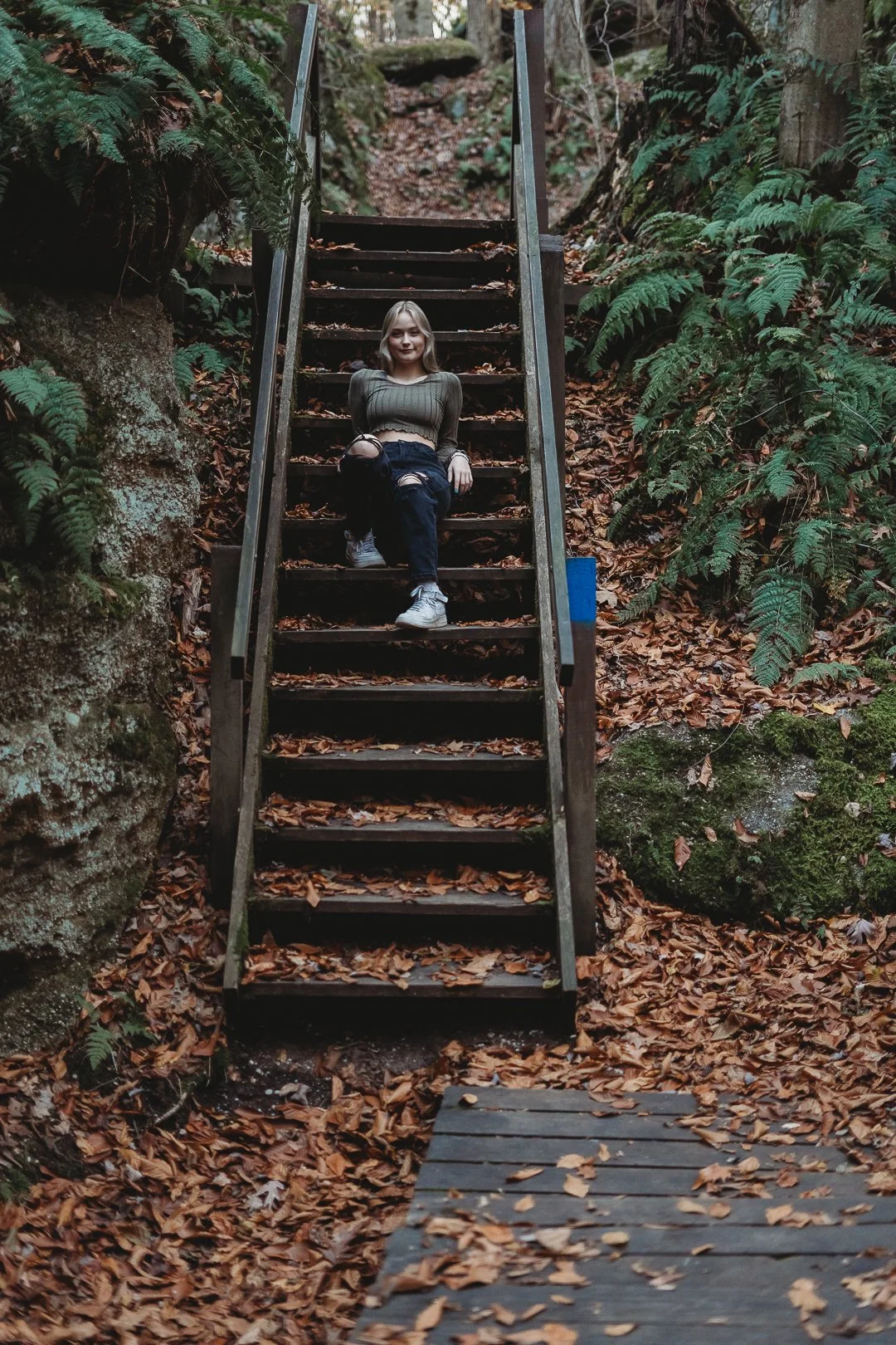 Girl sitting on wooden steps in a forest area, surrounded by ferns and fallen leaves.