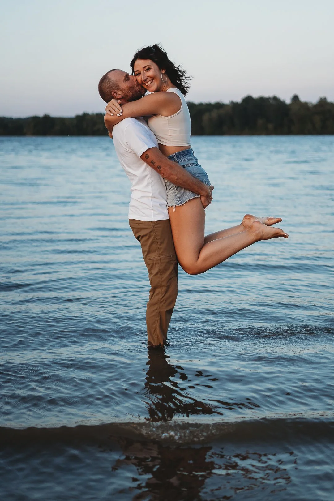 A couple embracing in a lake, with the man kissing the woman's cheek as he lifts her. The woman is smiling, wearing a white tank top and denim shorts. The man is in white t-shirt and tan pants, and the background shows trees and sky.