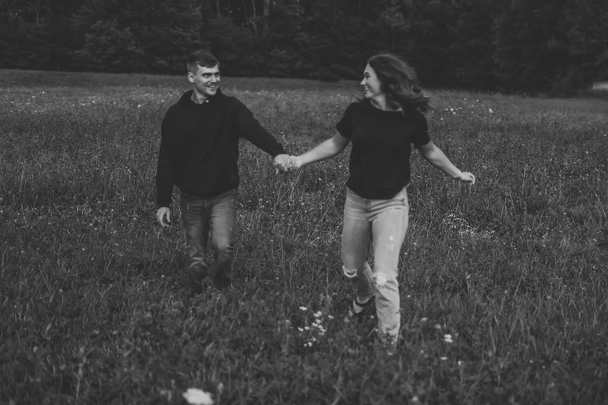 A couple holding hands while running through a grassy field with trees in the background, in black and white.