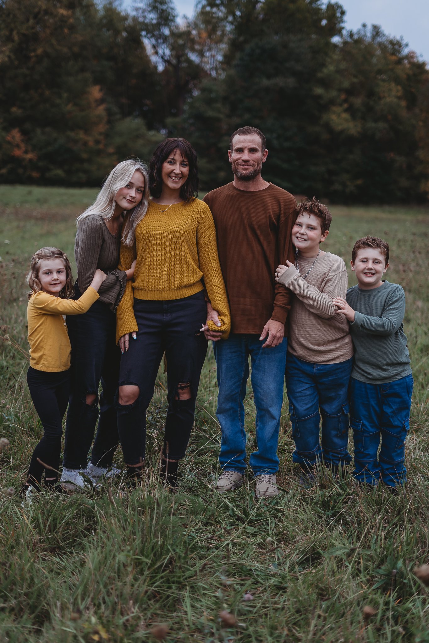 Family standing in a grassy field with trees in the background, wearing casual fall attire in neutral and earth tones; parents with three children.