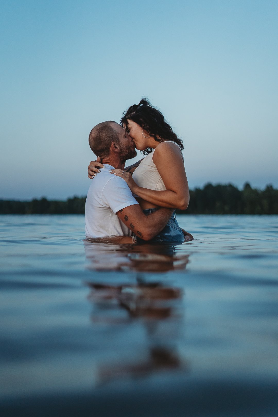 A couple kissing while standing in a lake at sunset, with trees in the background.