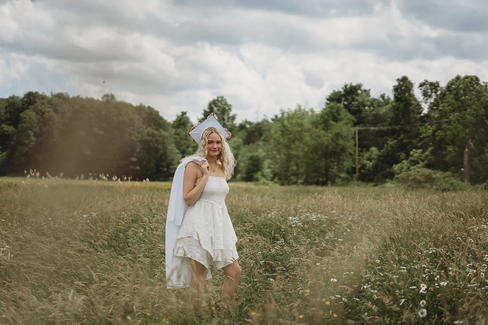 Young woman in a white dress and graduation cap standing in a field, holding a gown over her shoulder, surrounded by tall grass and trees in the background on a cloudy day.