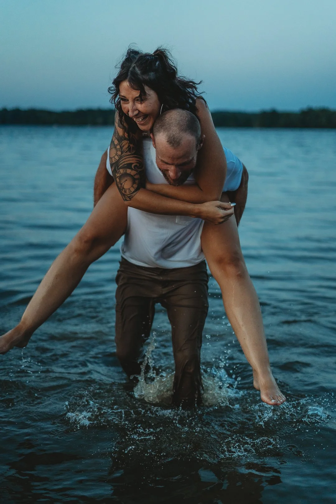 A joyful couple in a playful pose in a lake, with the man giving the woman a piggyback ride. The woman has a tattoo on her arm, and they are splashing in the water.