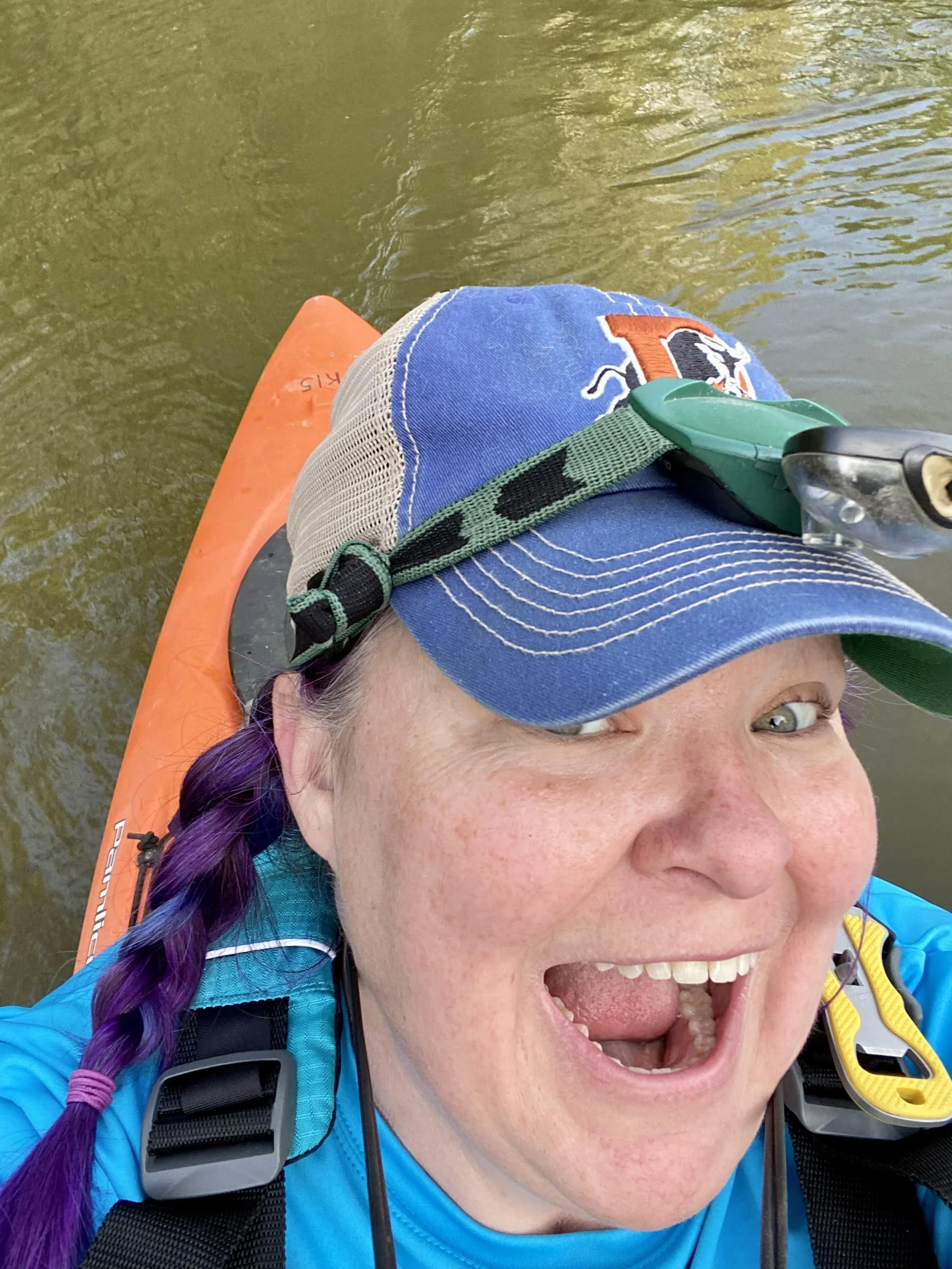 Woman smiling on a boat, wearing a baseball cap and life jacket, with a calm body of water and a canoe in the background.