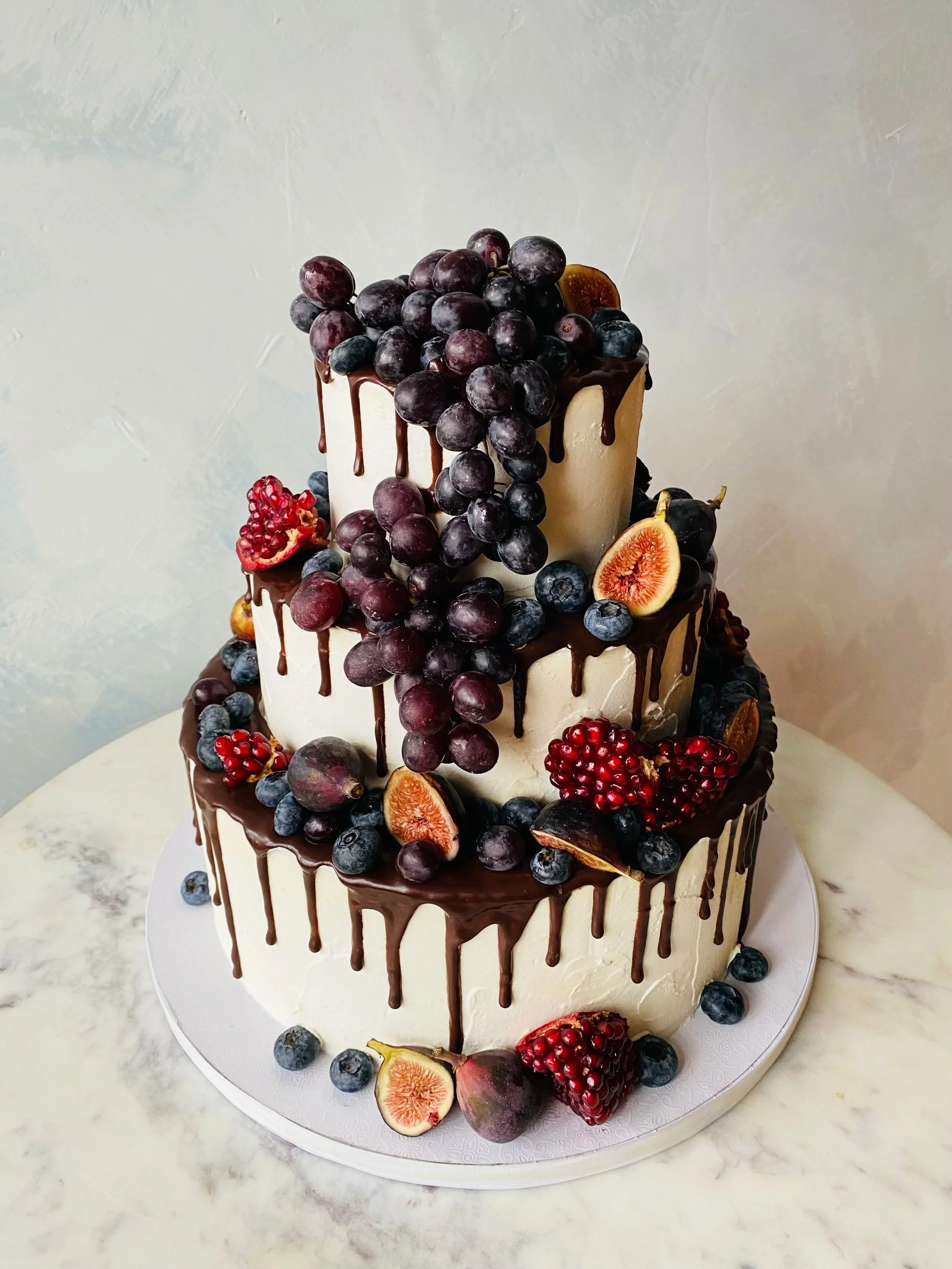 Three-tiered cake with white frosting, topped with fresh grapes, blueberries, figs, and drizzled with chocolate.