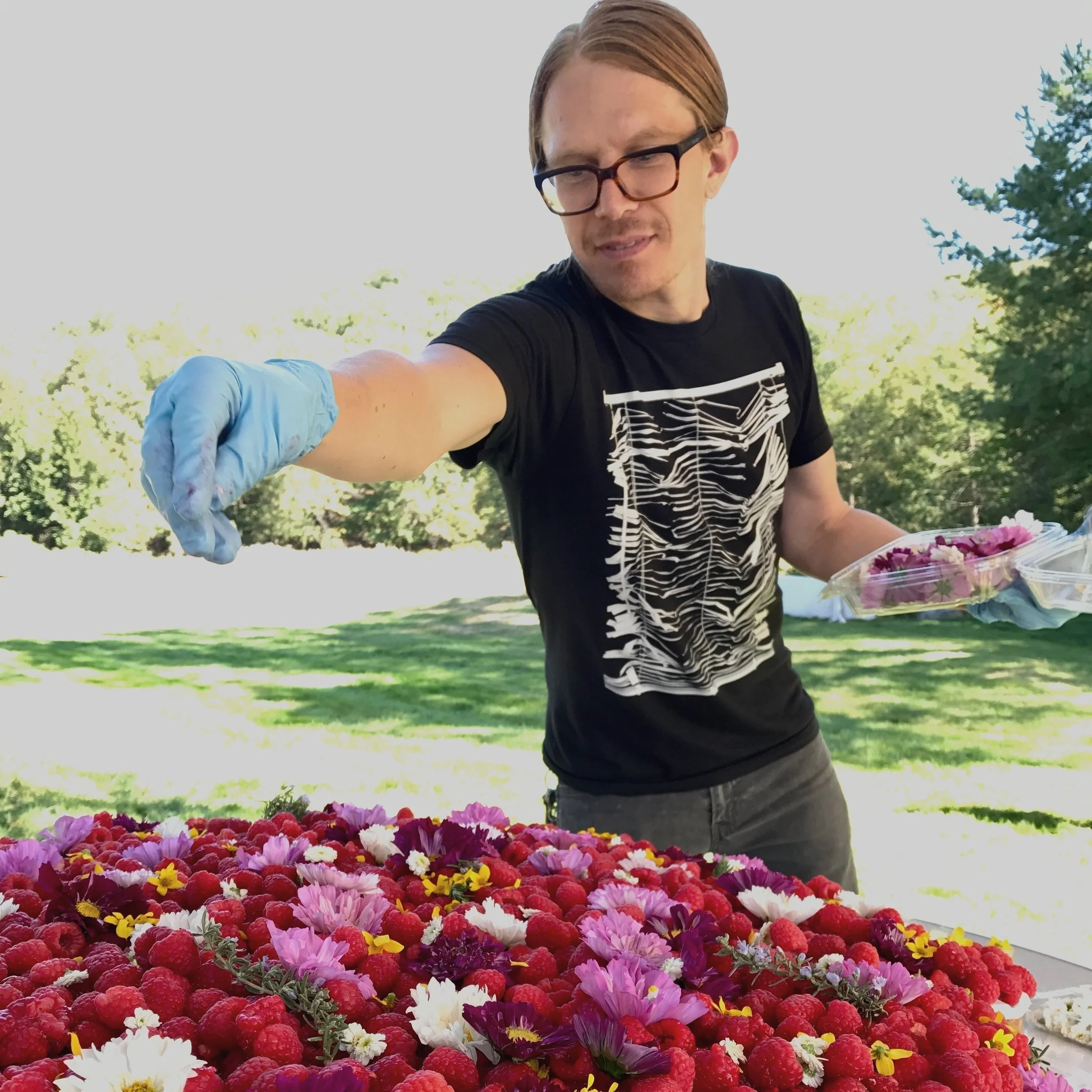A man with glasses wearing a black t-shirt and blue gloves stands outdoors, reaching out over a table filled with colorful flowers and berries, with a background of green trees and clear sky.