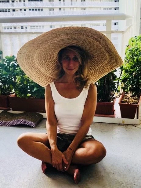 Woman sitting cross-legged on a balcony or patio, wearing a large straw hat, white tank top, and black shorts. Potted plants and city buildings are in the background.