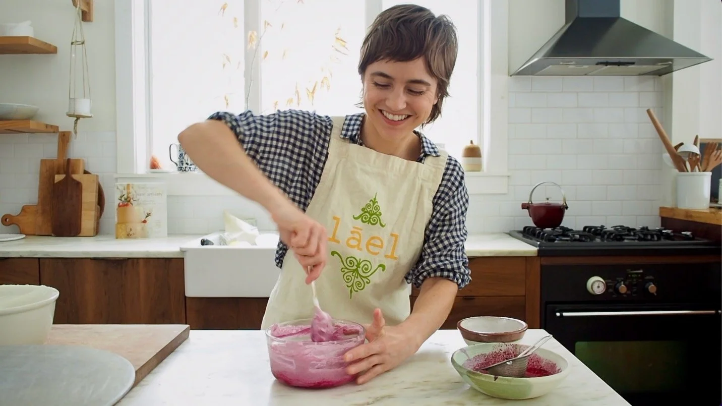 A smiling woman with short brown hair wearing a patterned shirt and an apron stirring a pink mixture in a glass bowl in a kitchen.