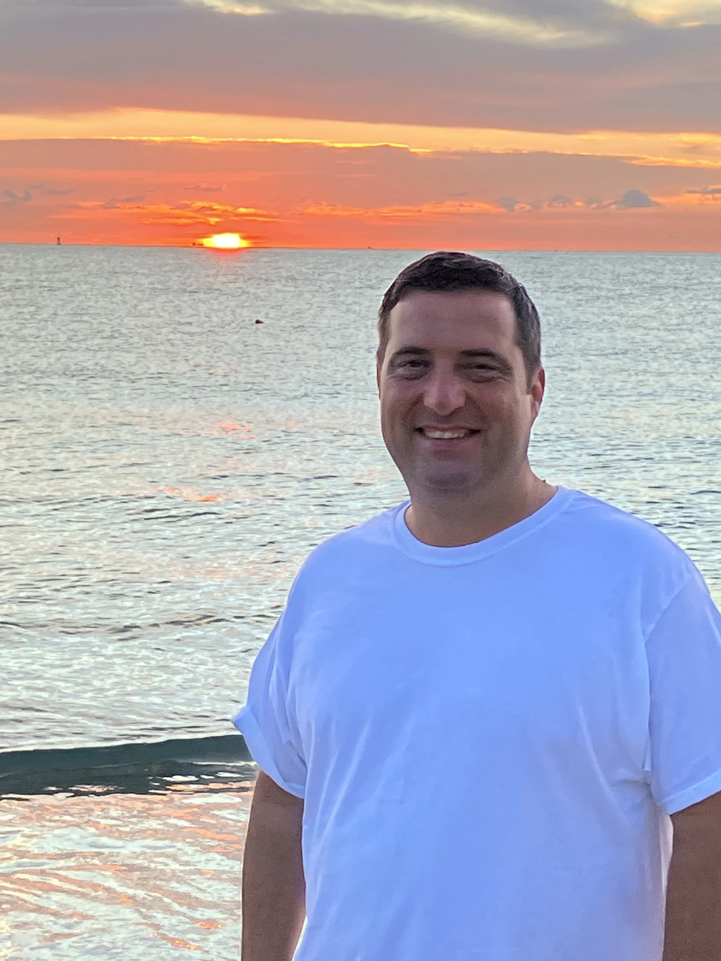 A smiling man in a white t-shirt standing in front of a body of water at sunset.