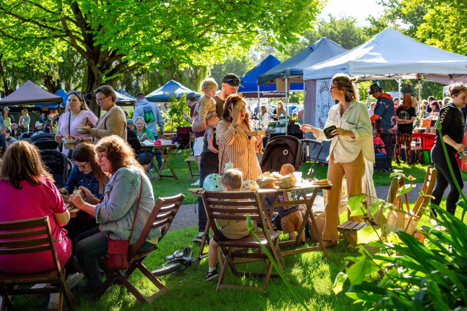People enjoying outdoor food fair with tents and trees, some sitting at tables and others standing in line, during sunny day with green foliage.