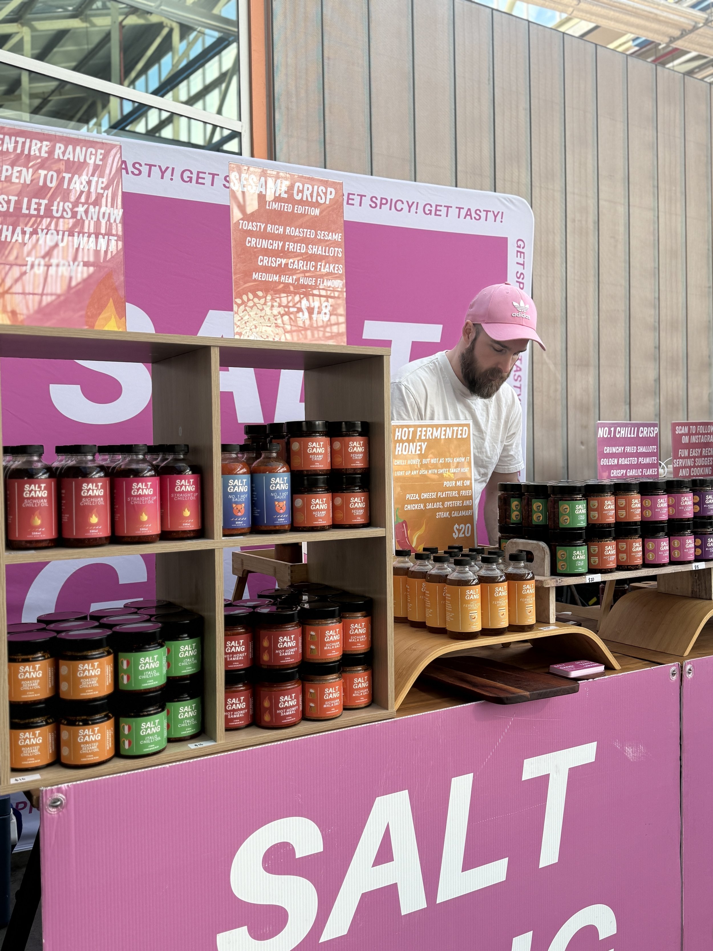 A man wearing a pink cap and white t-shirt standing behind a pink and wooden salt product display at an indoor event. The display features various jars of flavored salts with colorful labels and a large pink background banner with promotional signs for sesame crisp and hot fermented honey.