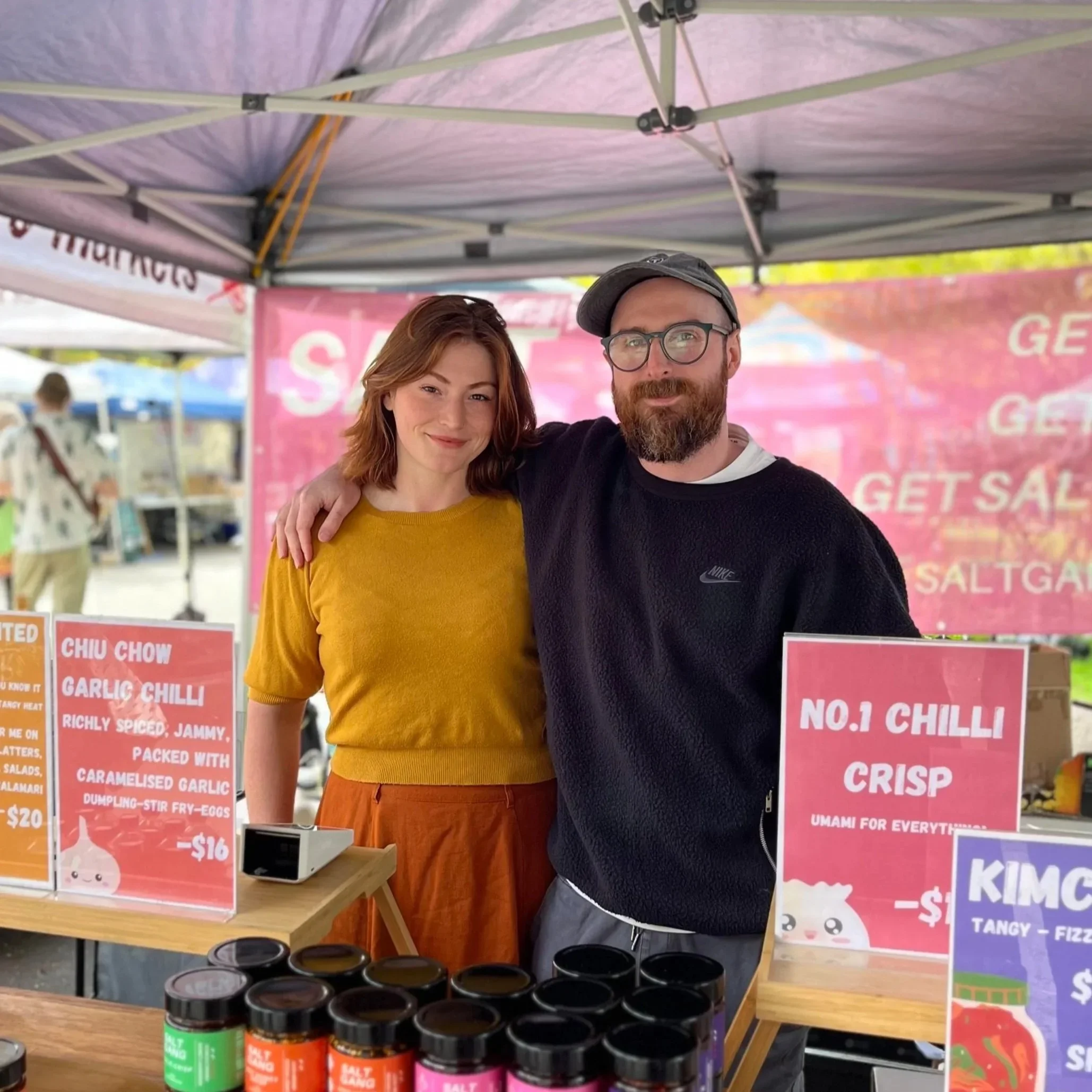 People shopping at an outdoor market with white tents under a sunny sky.