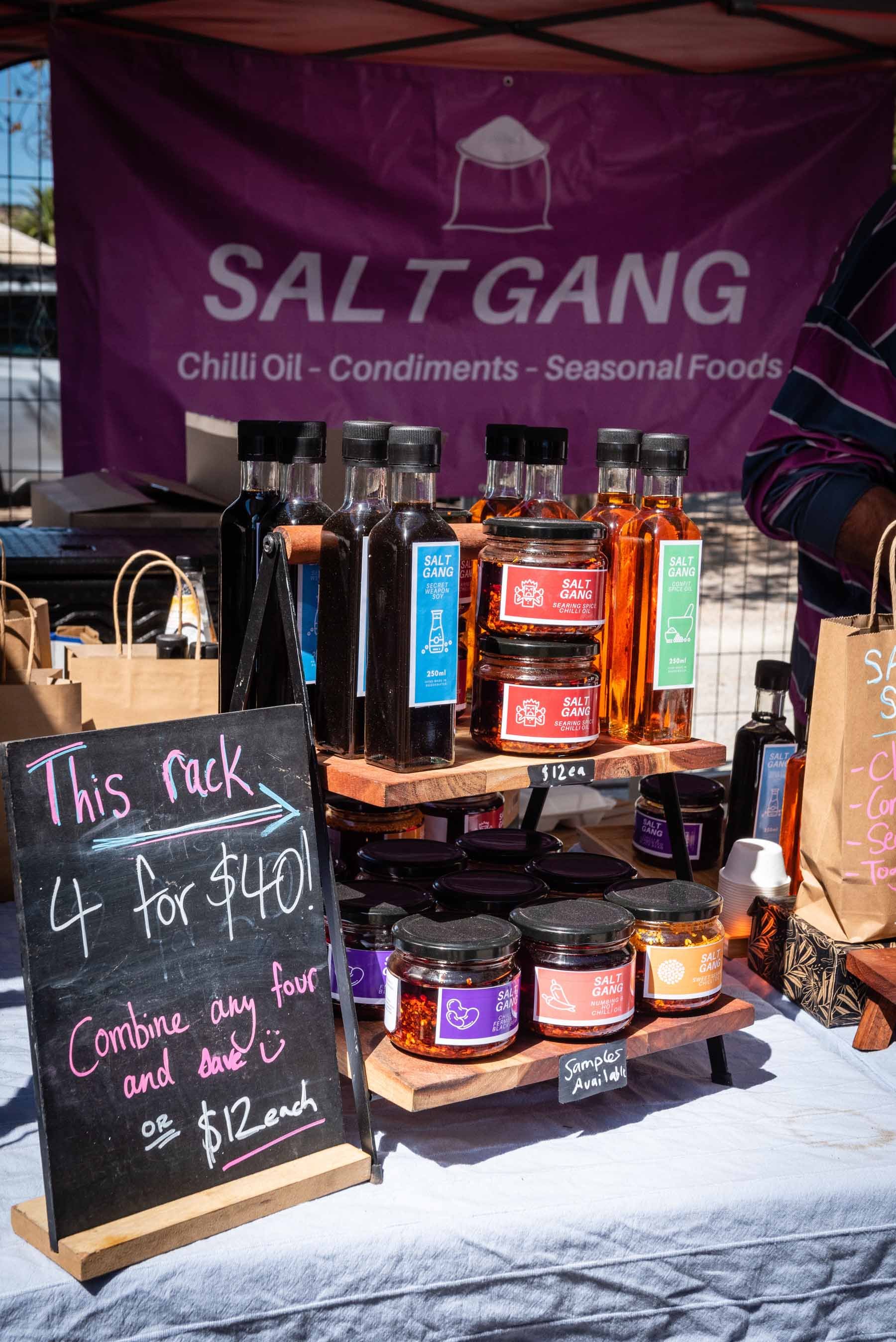 A display stand at a market stall with various jars and bottles of chili oil, condiments, and seasonal foods. A sign advertises four jars for $40 or individual jars for $12 each.